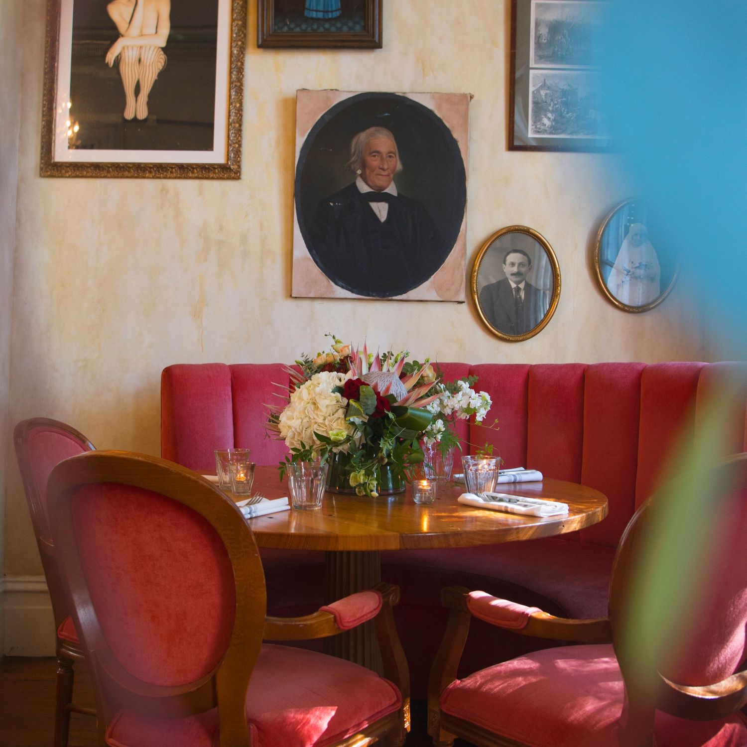 Elegant restaurant booth with red velvet seating, round wooden table, floral centerpiece, and vintage portrait paintings on textured walls.