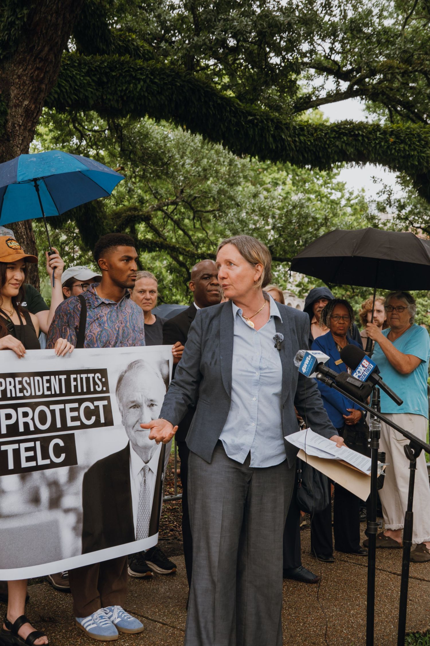 Woman speaking at outdoor press conference with crowd holding President Fitts Protect TELC protest sign.