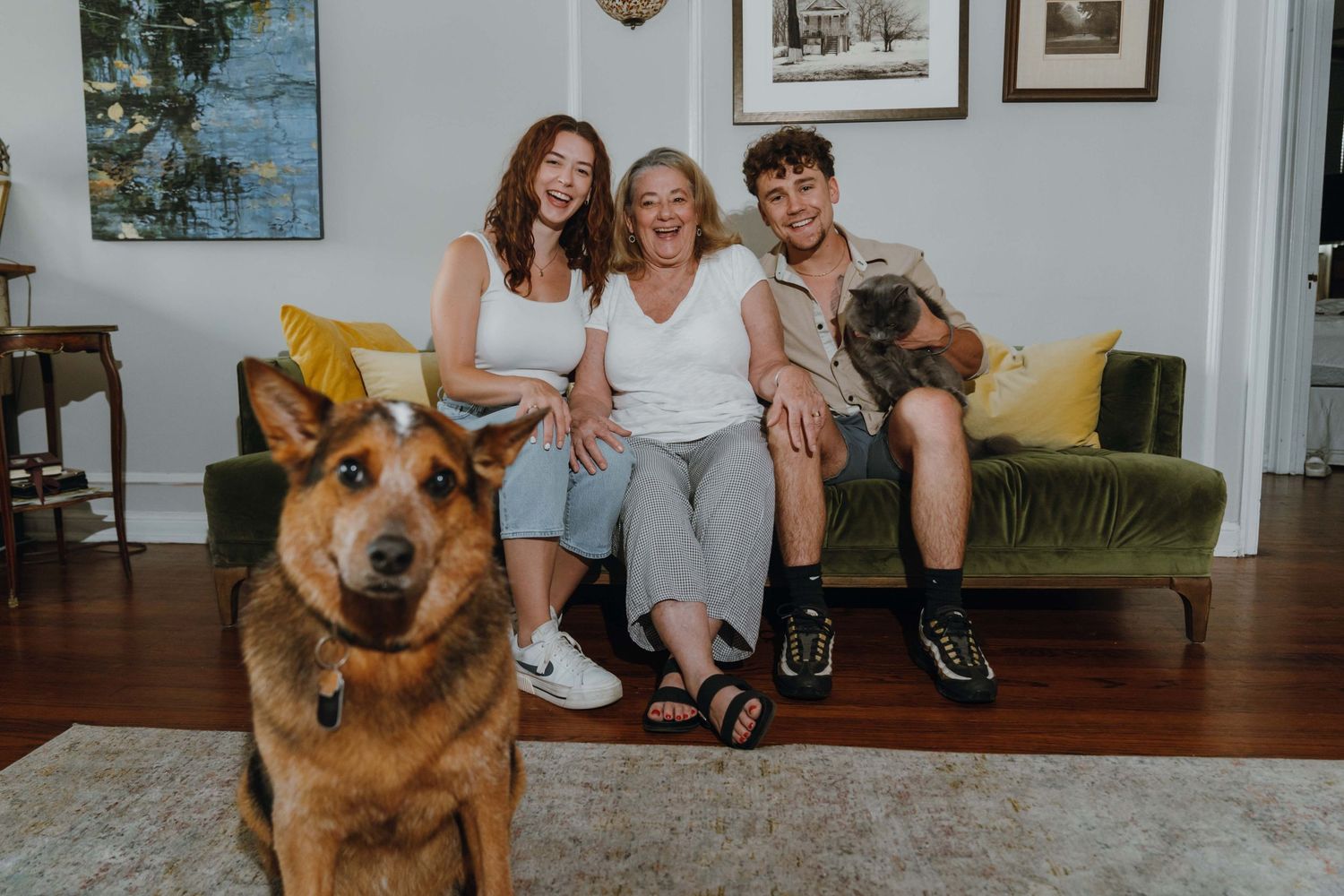 Three people smile on a green velvet sofa with a pug while a German Shepherd poses in the foreground of a bright living room.