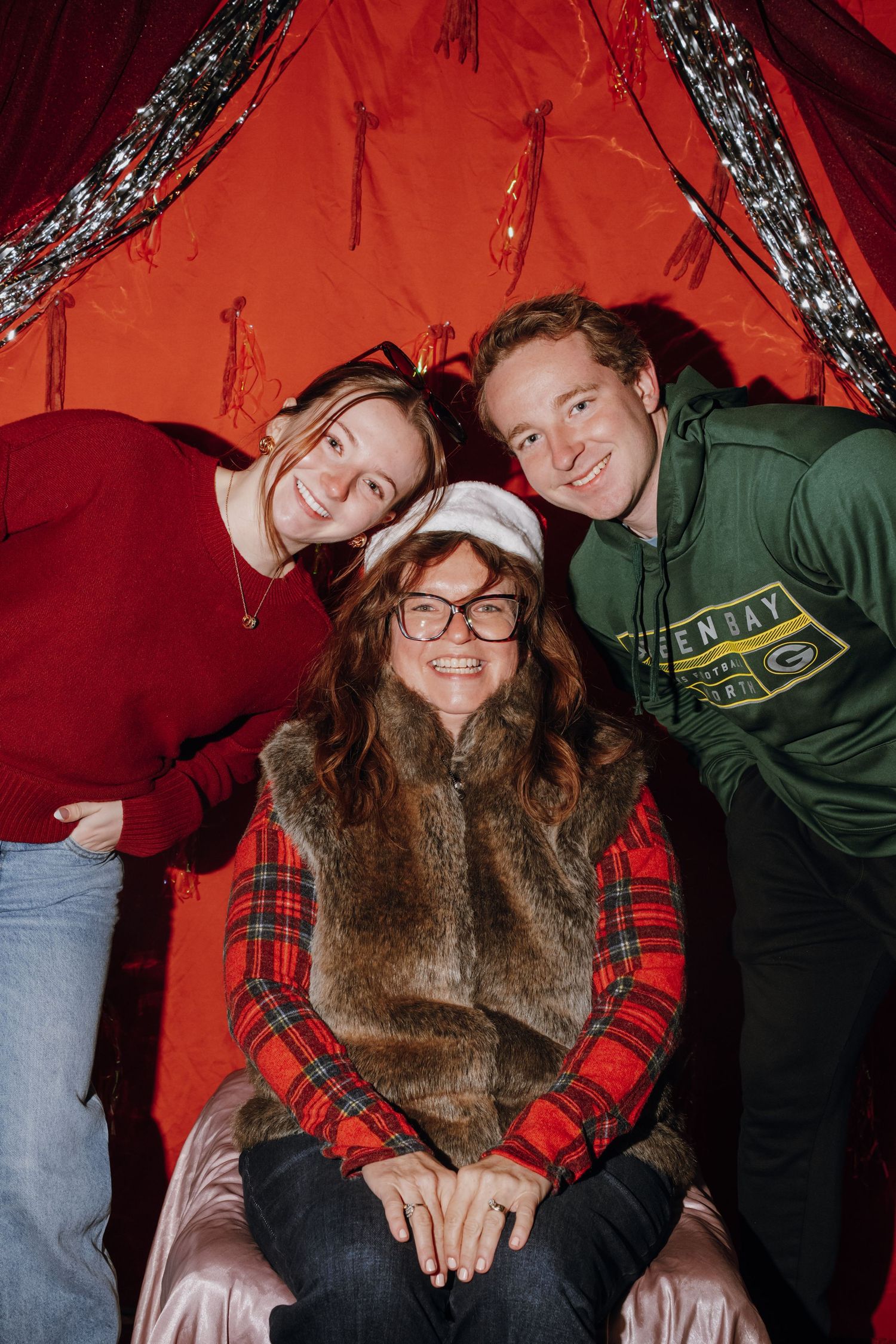 Three smiling people pose together in front of a festive red backdrop with holiday lights and silver tinsel.