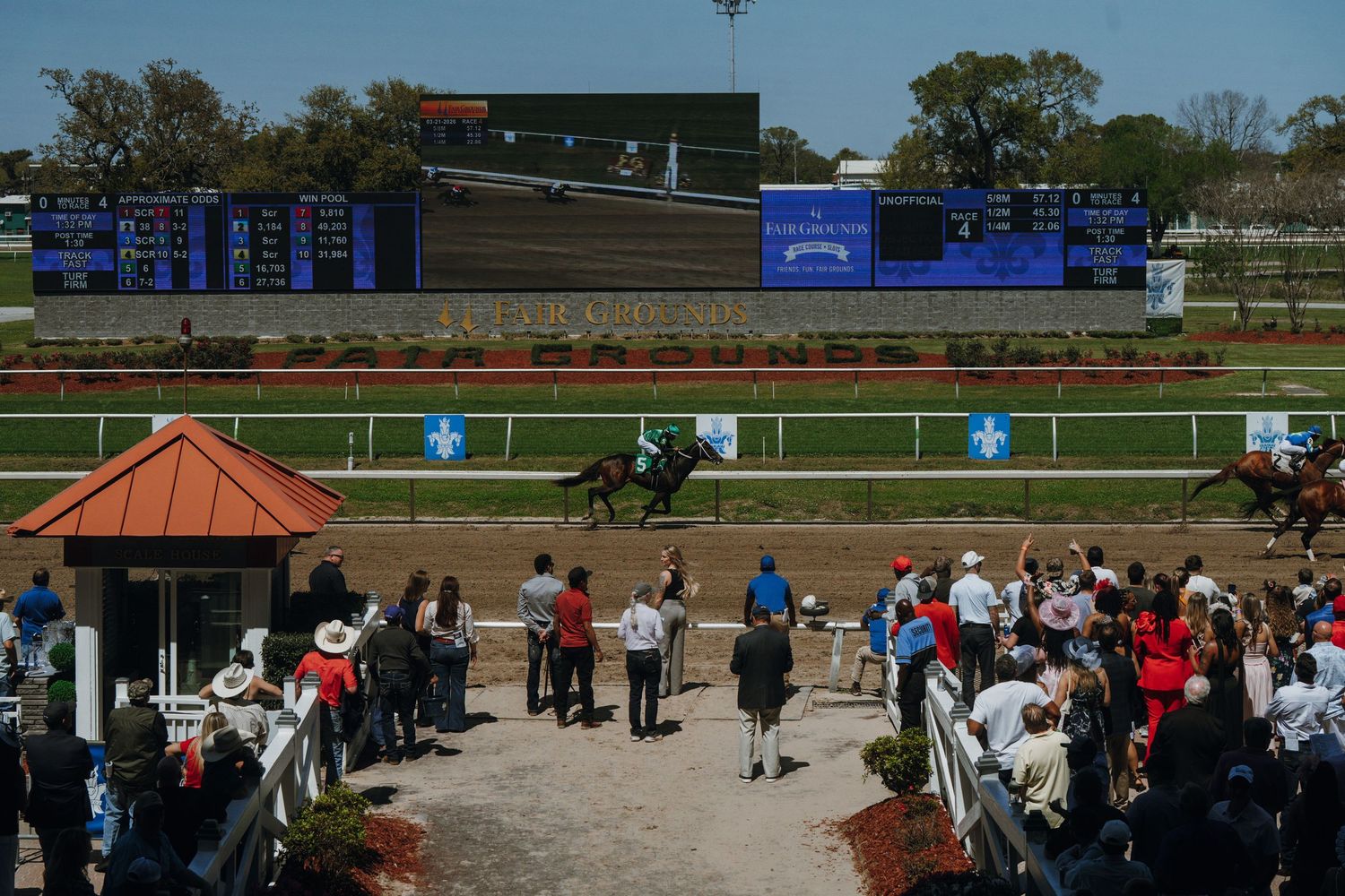 Horses racing on a dirt track at a crowded thoroughbred horse racing venue with a large scoreboard display.