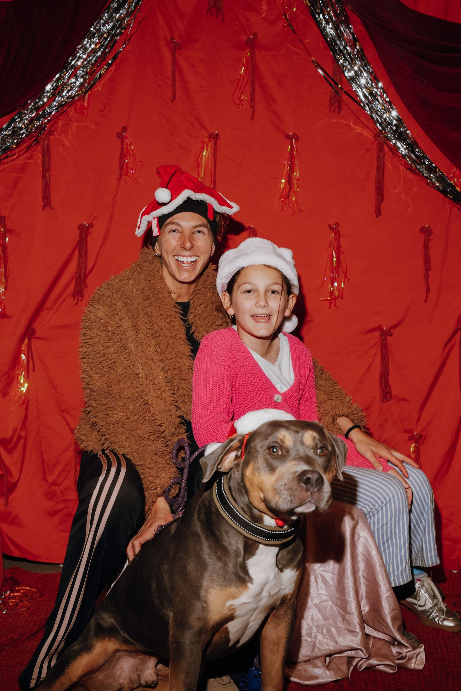 Woman and child in Santa hats pose with a Rottweiler dog in front of festive red Christmas backdrop.
