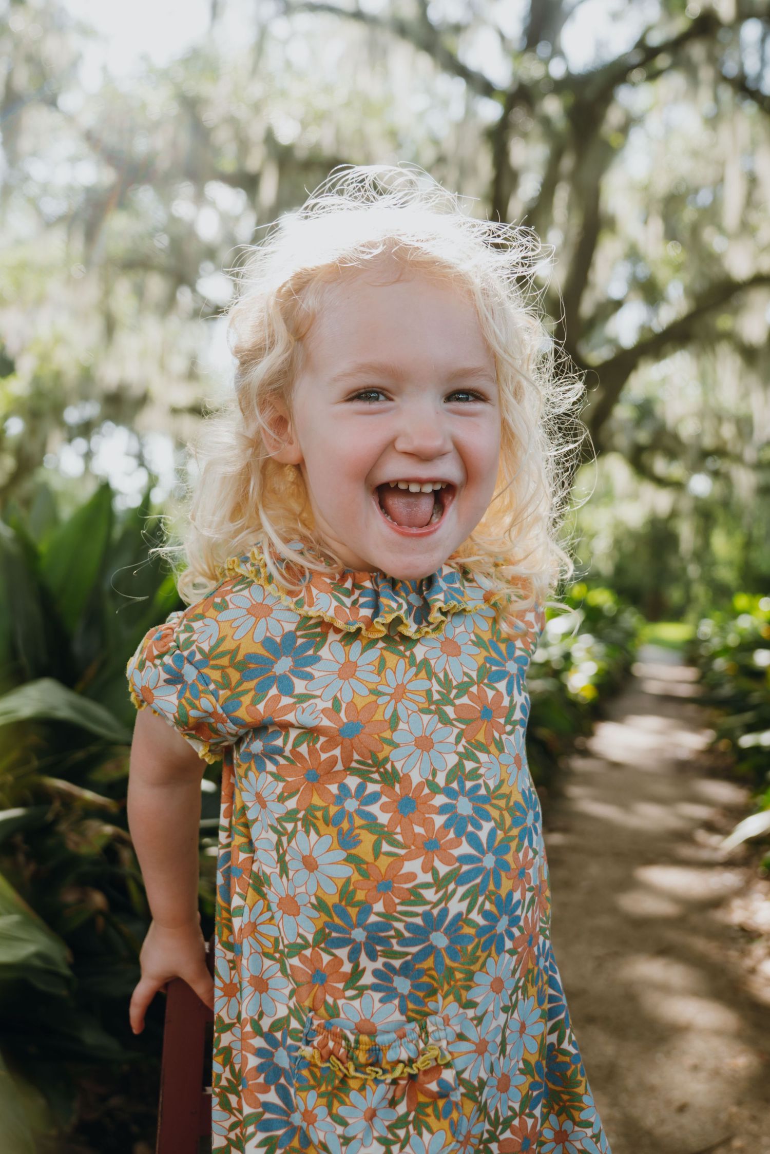 Laughing blonde toddler girl in floral dress standing on a garden path surrounded by lush green trees.