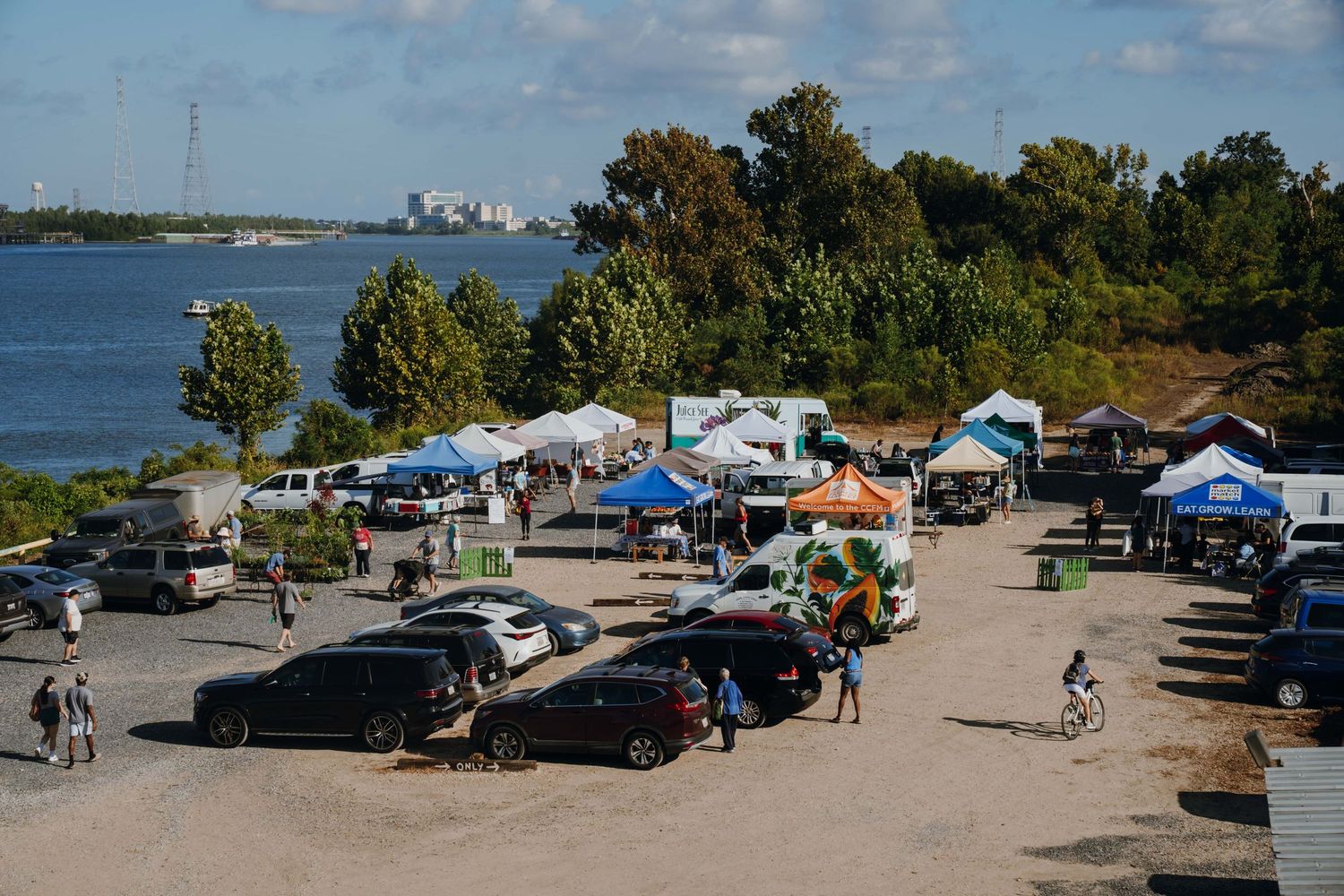Aerial view of a waterfront farmers market with vendor tents and cars.