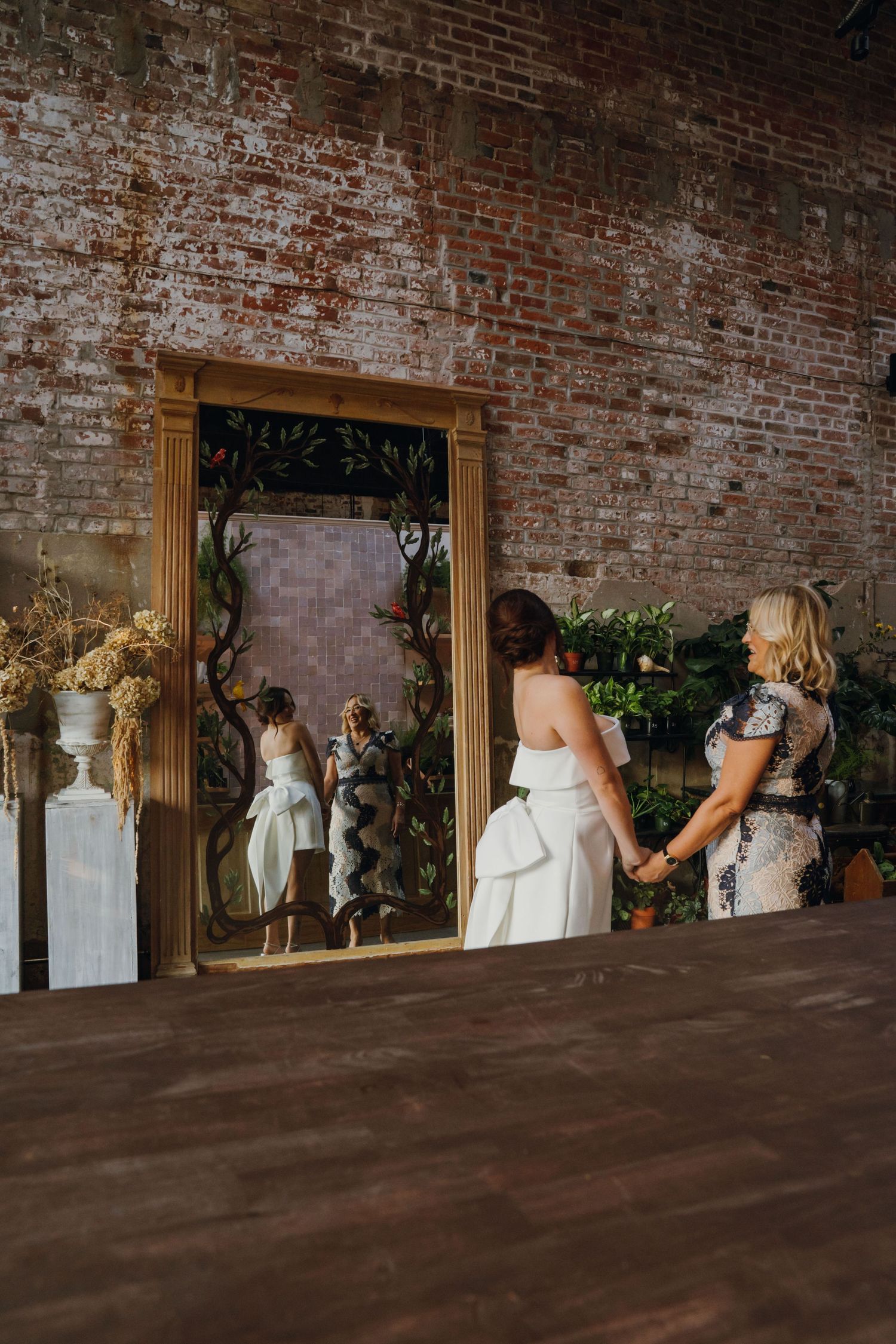 Bride in white mini dress views ornate gold mirror with floral details at rustic brick wedding venue.