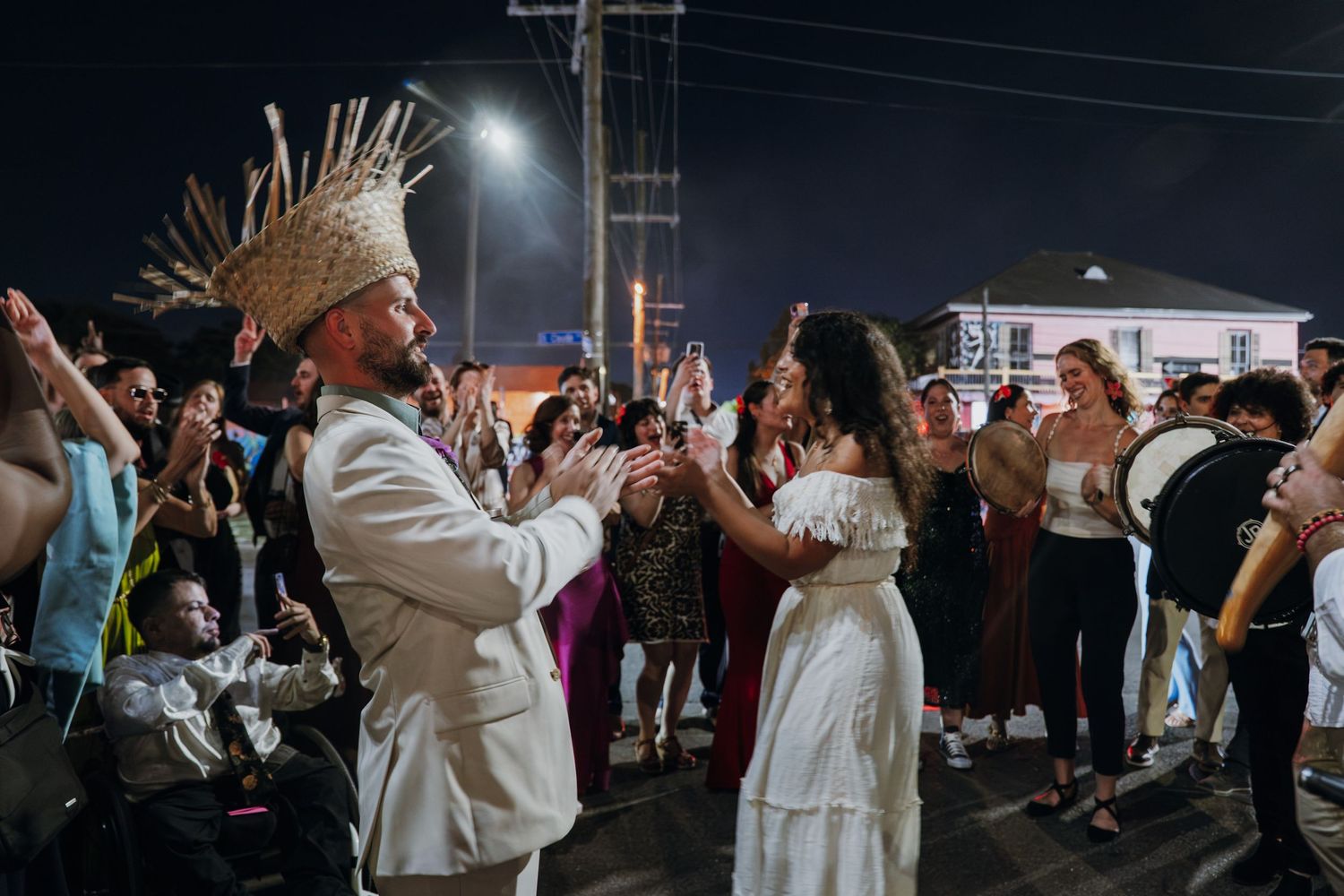 Bride and groom in white outfits dancing joyfully with cheering wedding guests on street at night.