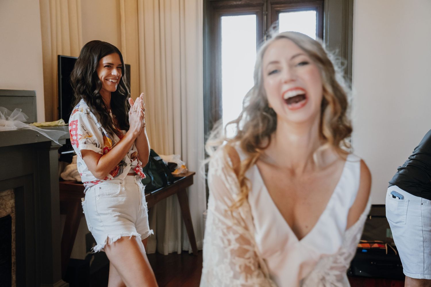 Joyful bride laughing with excited bridesmaid clapping during wedding morning preparations.