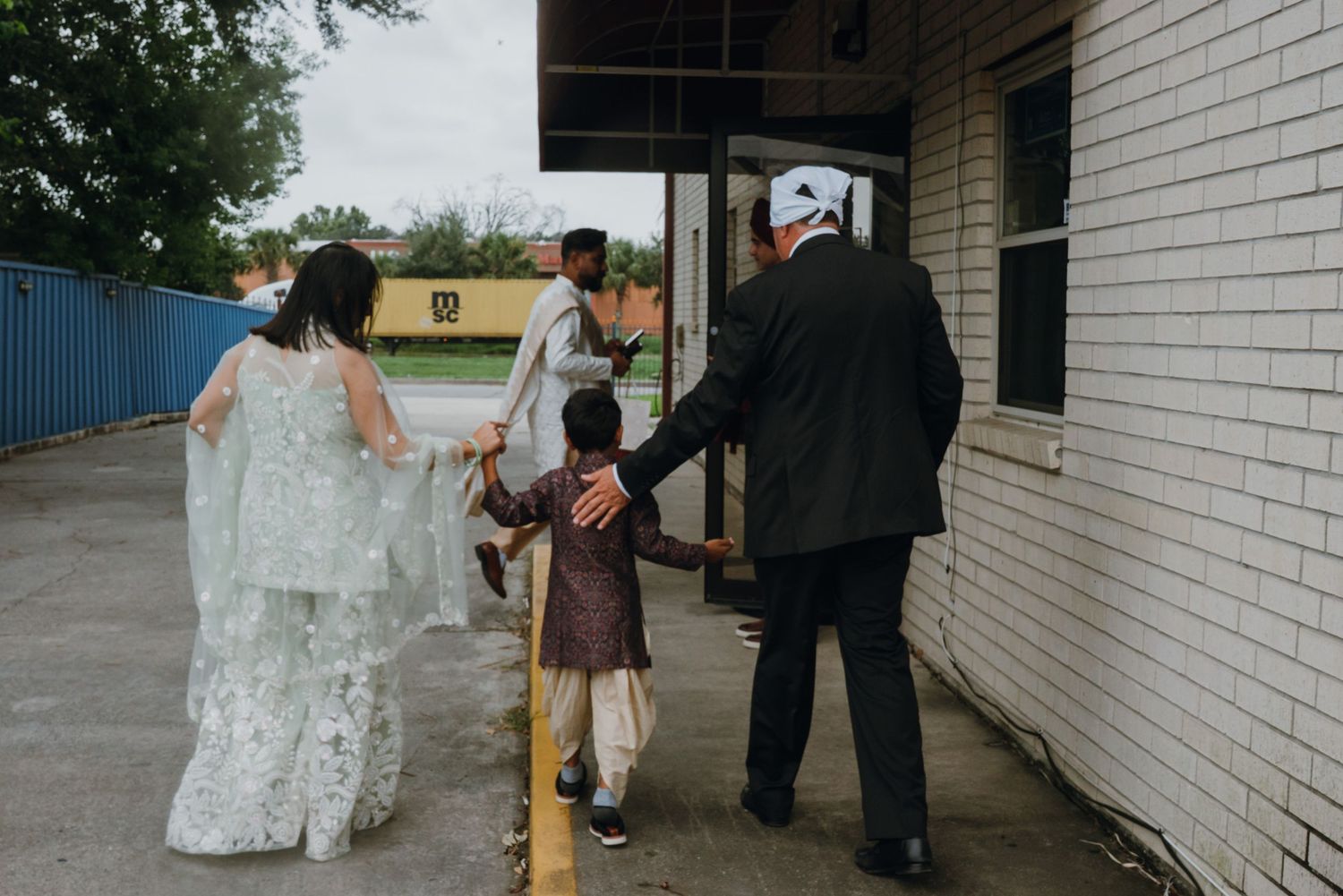 Wedding party including child walks along building exterior toward South Asian wedding venue.