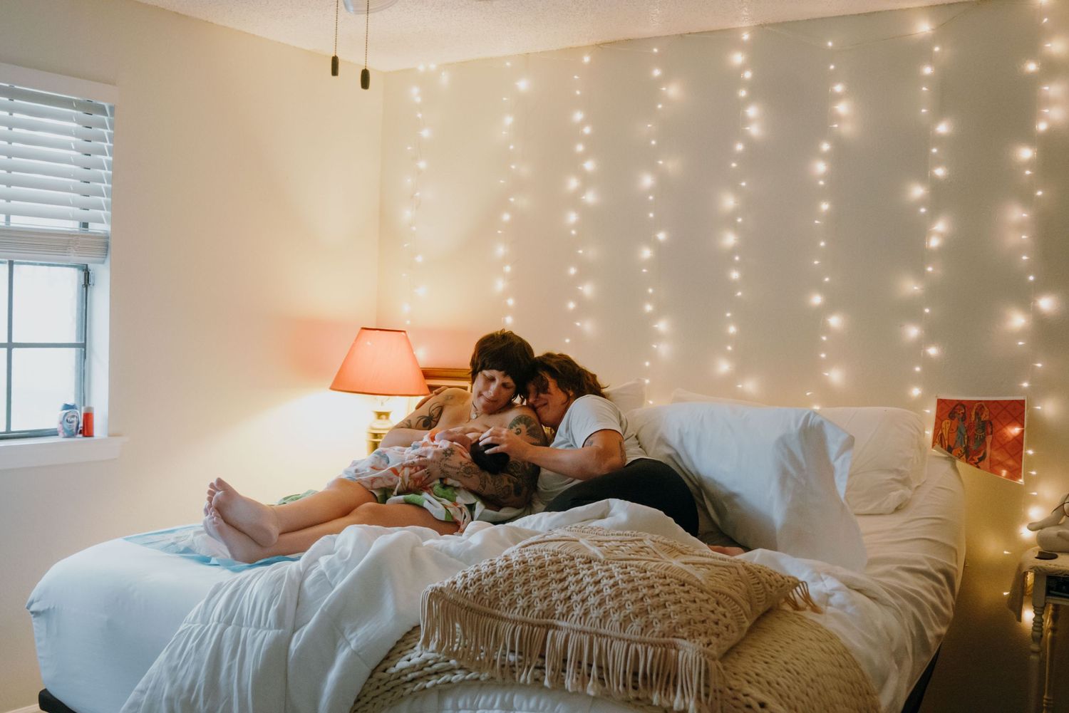 Two people sit together on a bed against a glowing fairy light wall holding a newborn.