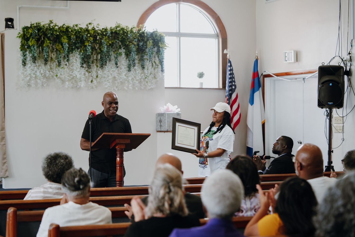 Two people presenting framed certificate award to audience in decorated church interior.