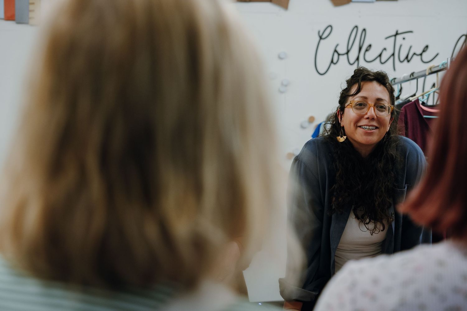 Curly-haired woman with glasses smiling warmly during a discussion inside a collective community space.