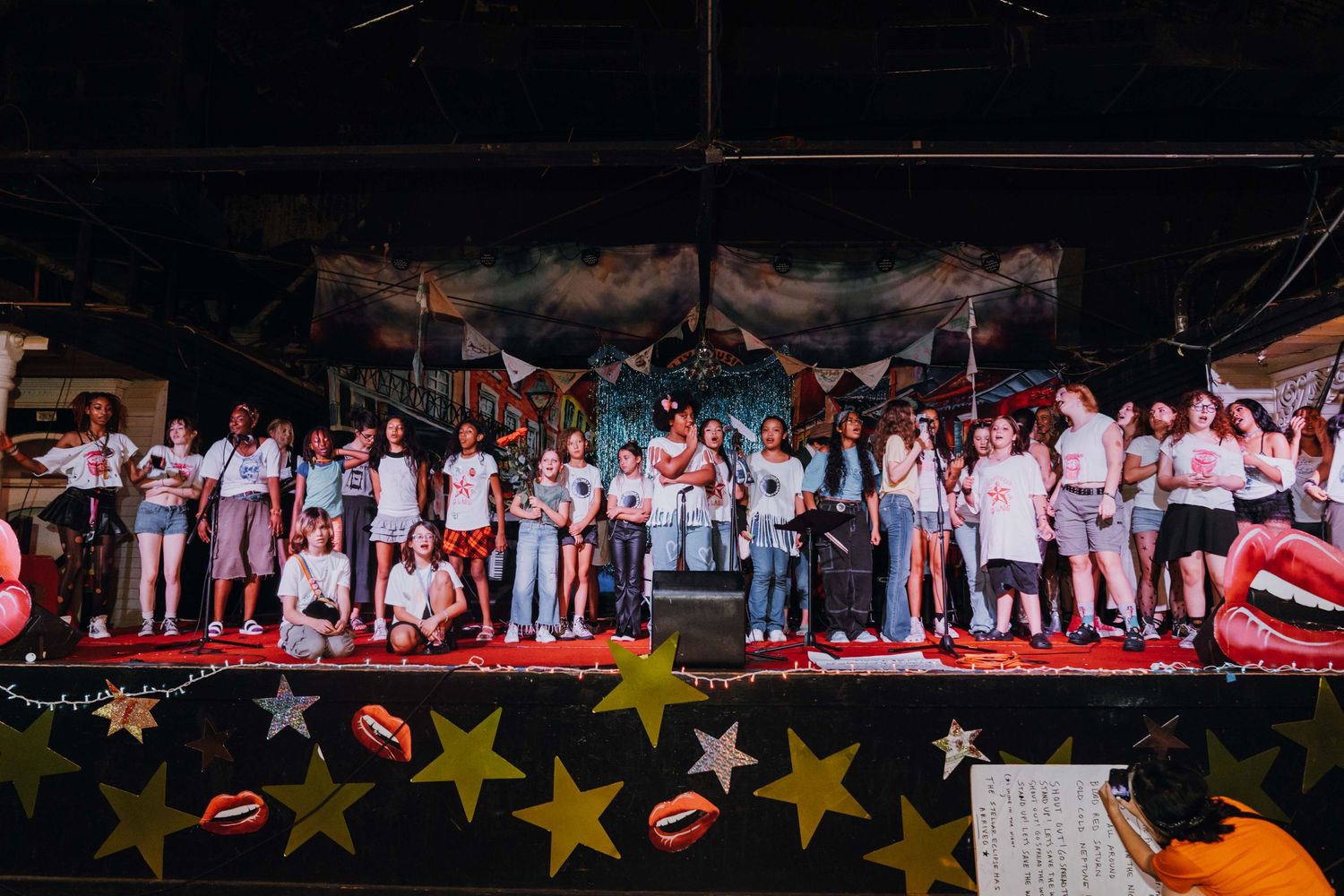 Large group of performers on a colorful stage decorated with stars and bunting at a live event.