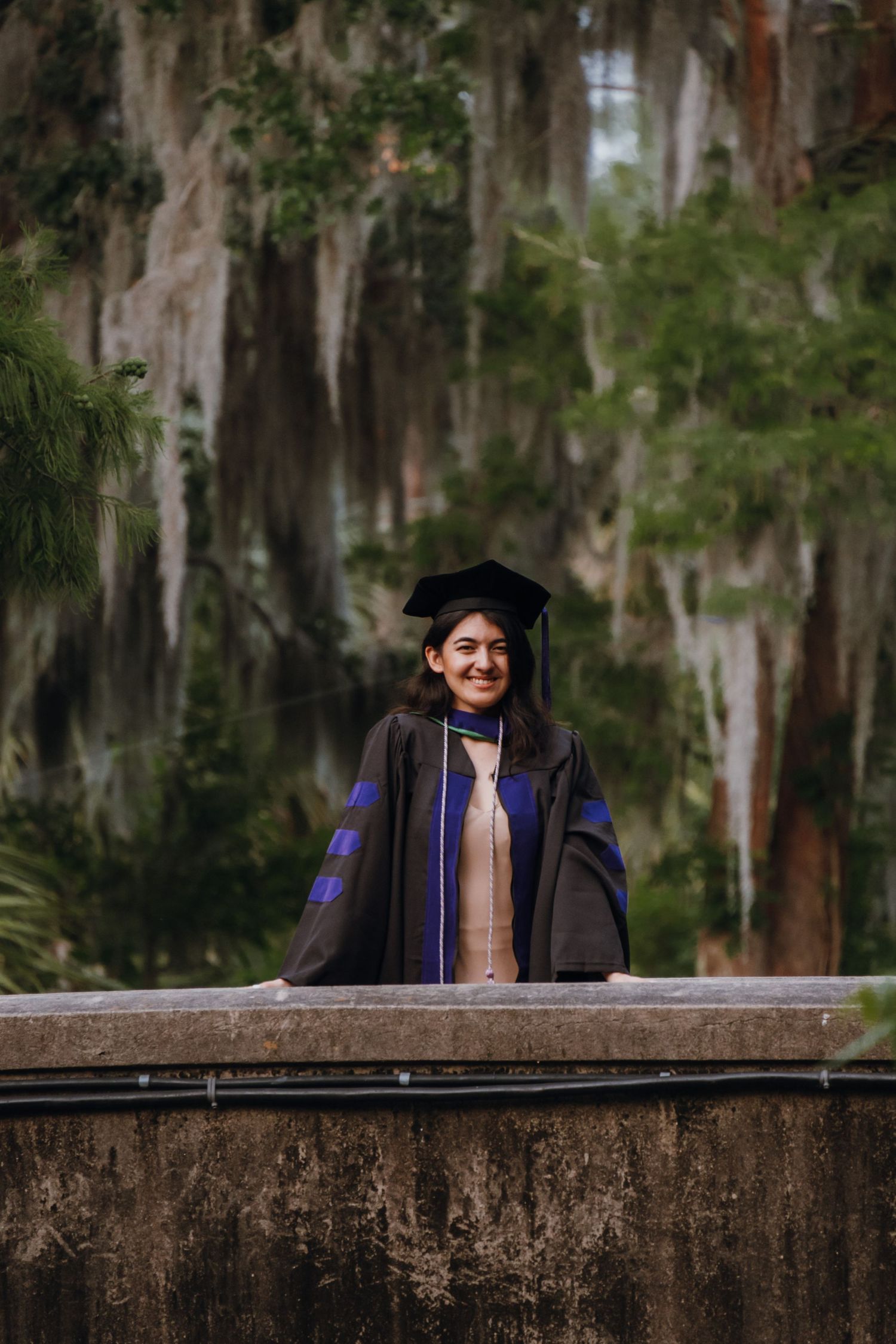 Smiling graduate in black and blue doctoral cap and gown standing before Spanish moss trees.