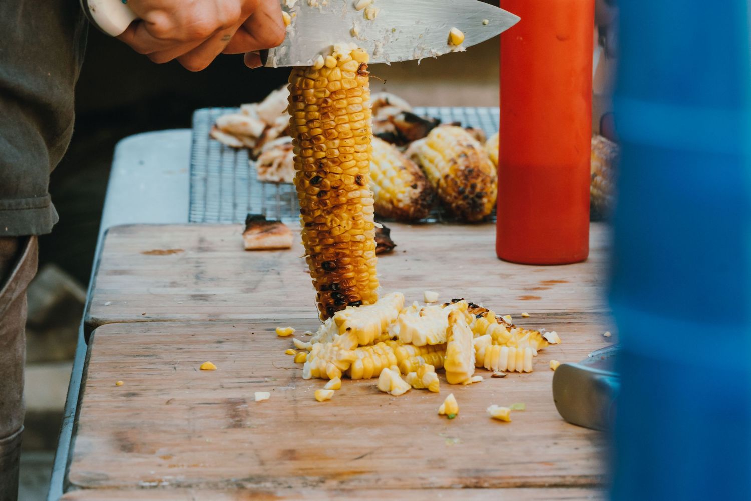 Person cutting grilled corn kernels off the cob on a wooden cutting board outdoors.