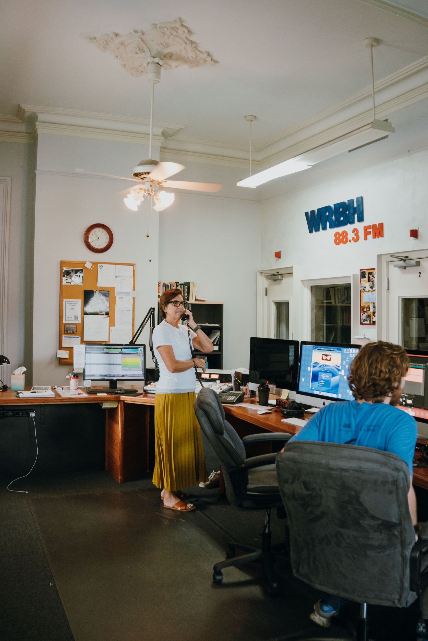 Two people working at desks in a WRBH 88.3 FM radio station office with computers and bulletin board.