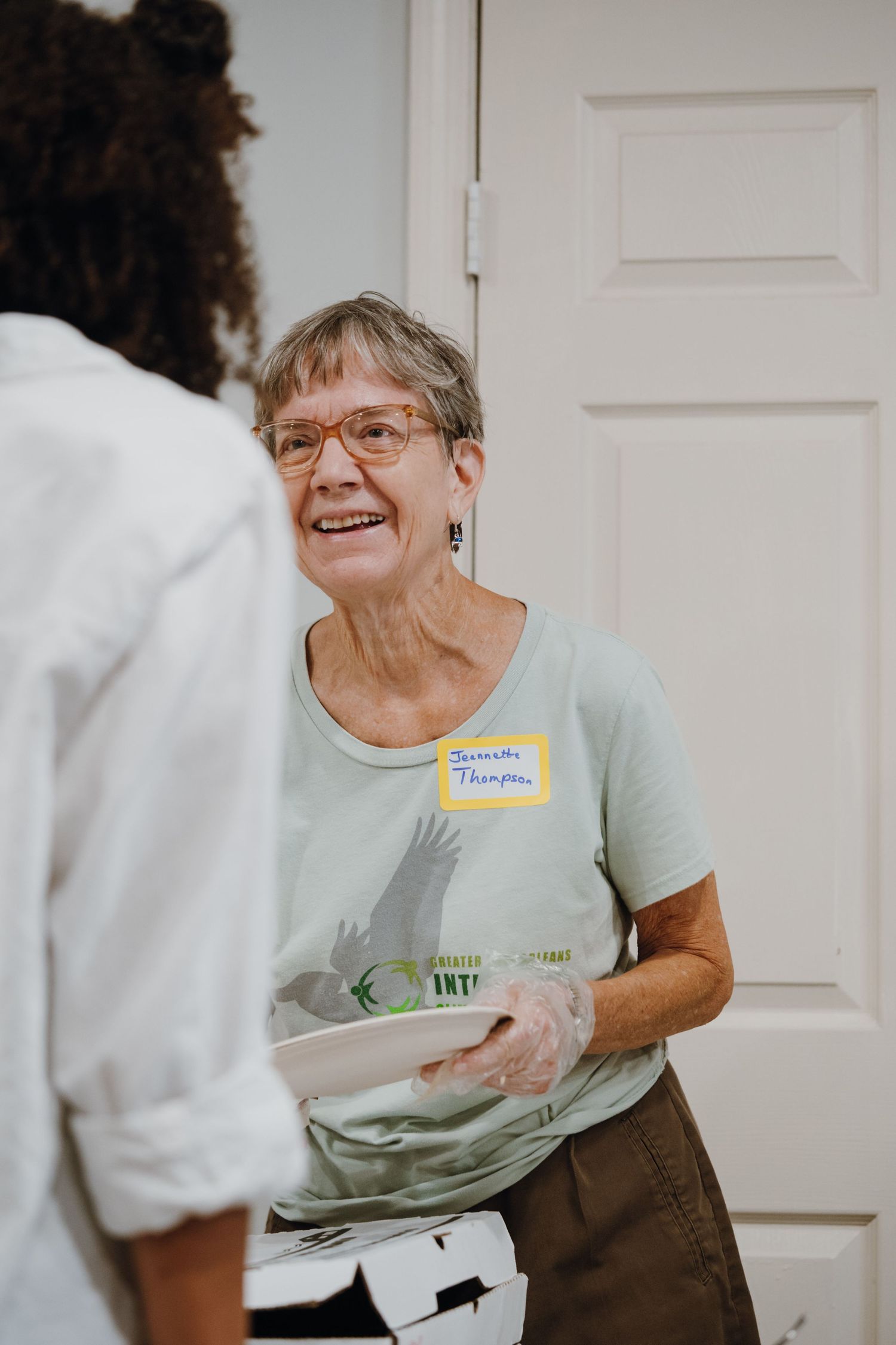 Smiling elderly volunteer named Jeanette Thompson wearing name tag serving food at community gathering.