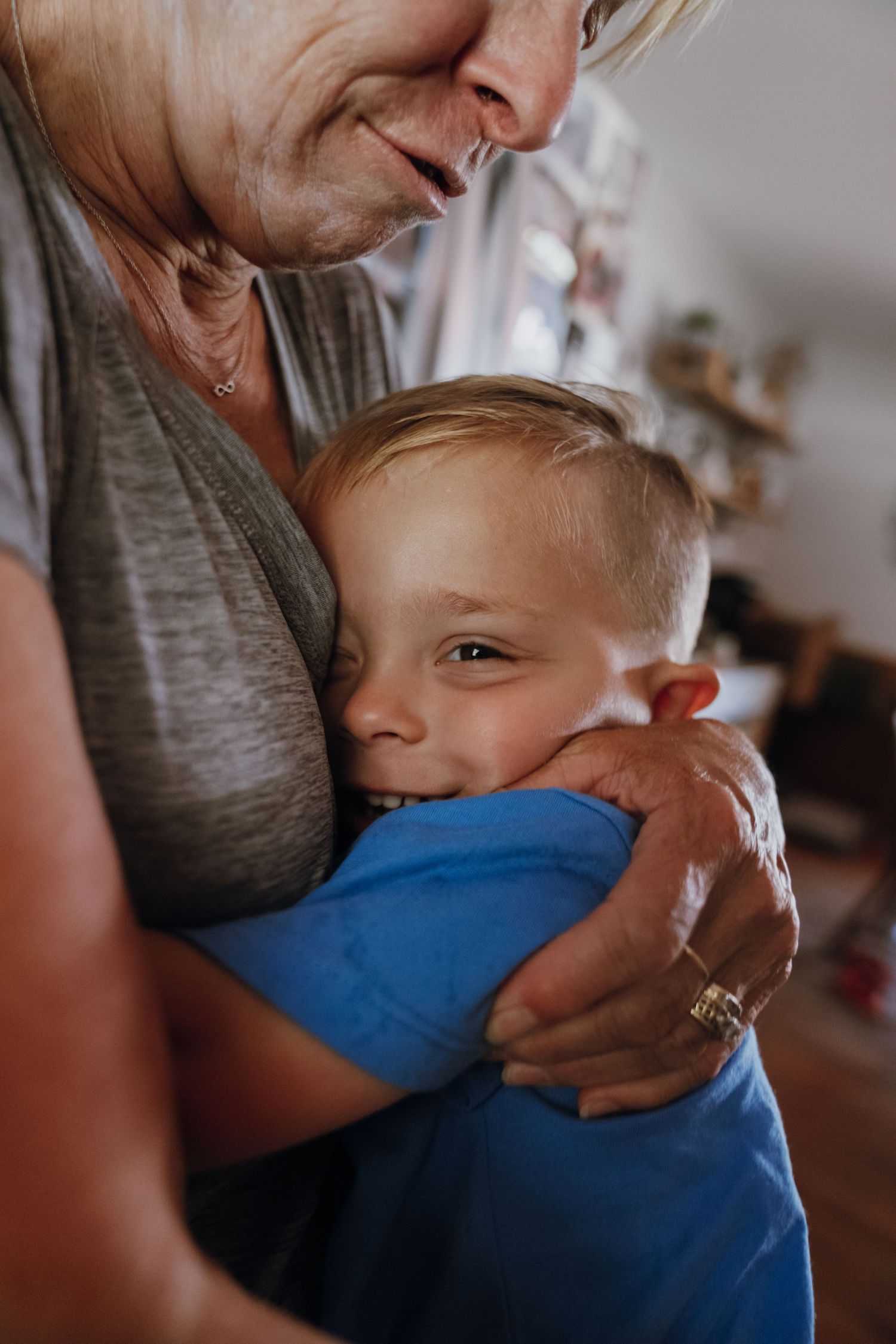 Grandmother smiling while hugging a young blonde boy in a blue shirt indoors.
