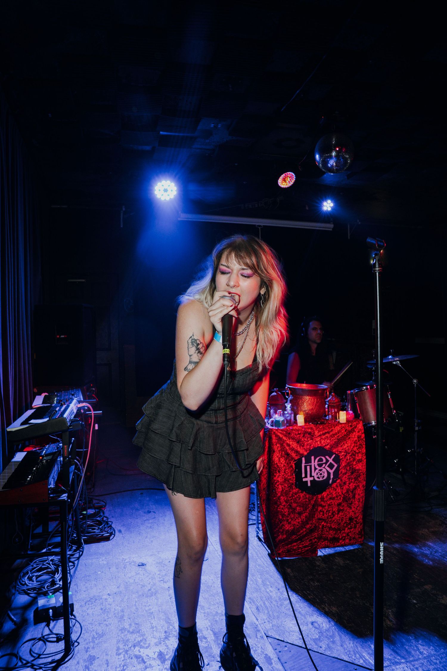 Female singer in black dress performing on stage under blue spotlights with drummer in background.