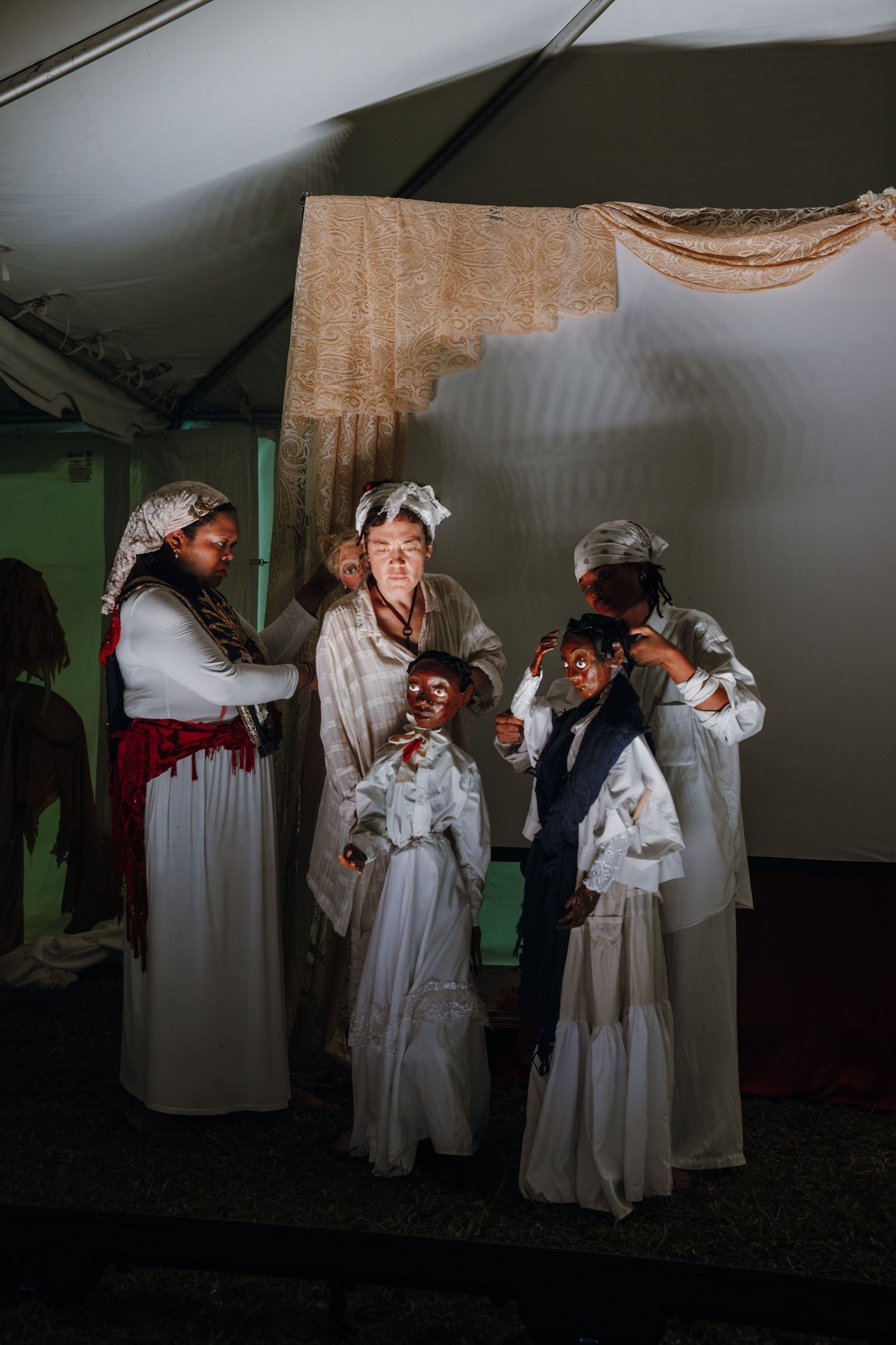 Group of performers in white robes and masks prepare backstage under a tent at night.
