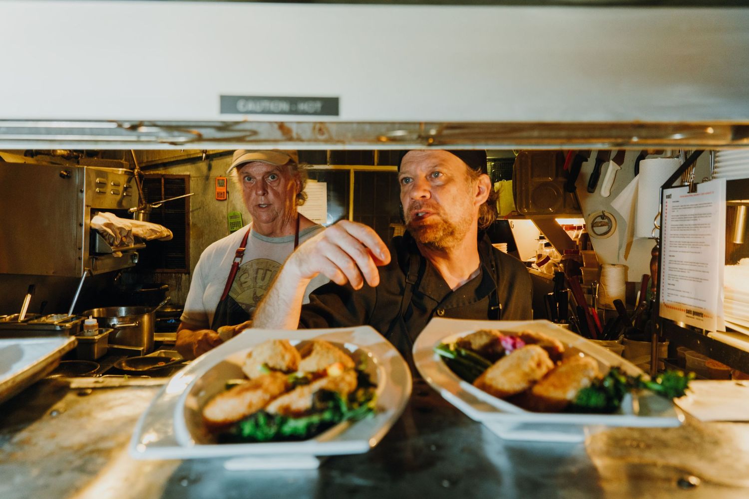 Two chefs plate gourmet dishes at a restaurant kitchen pass, carefully garnishing food under warm lighting.