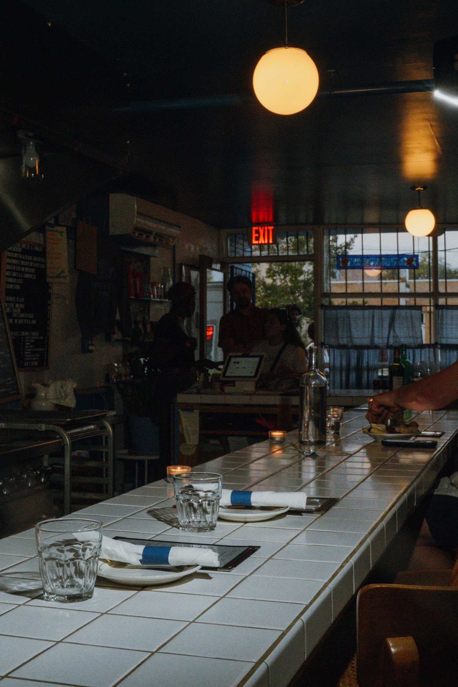 Dark restaurant interior with tiled bar counter, pendant lights, and place settings in the foreground.