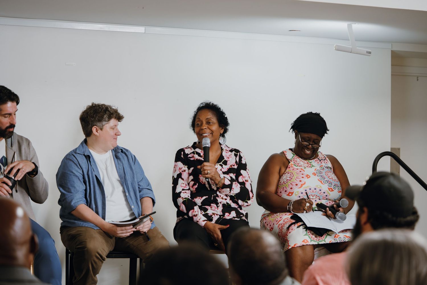 A diverse panel of four speakers seated in chairs, with a woman in a floral dress speaking into a microphone.