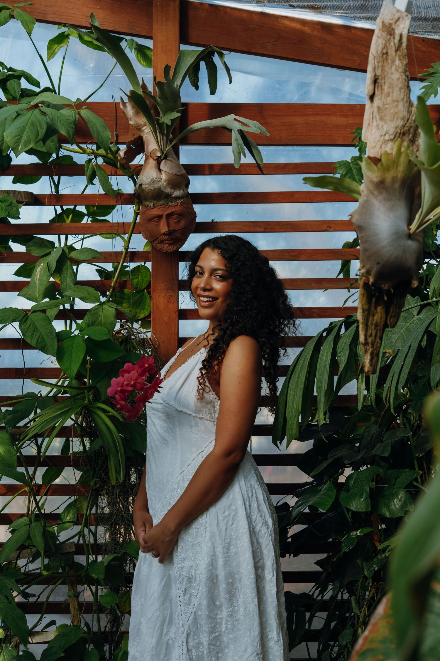 A smiling woman in a white dress stands among tropical plants holding a pink flower in a wooden greenhouse.
