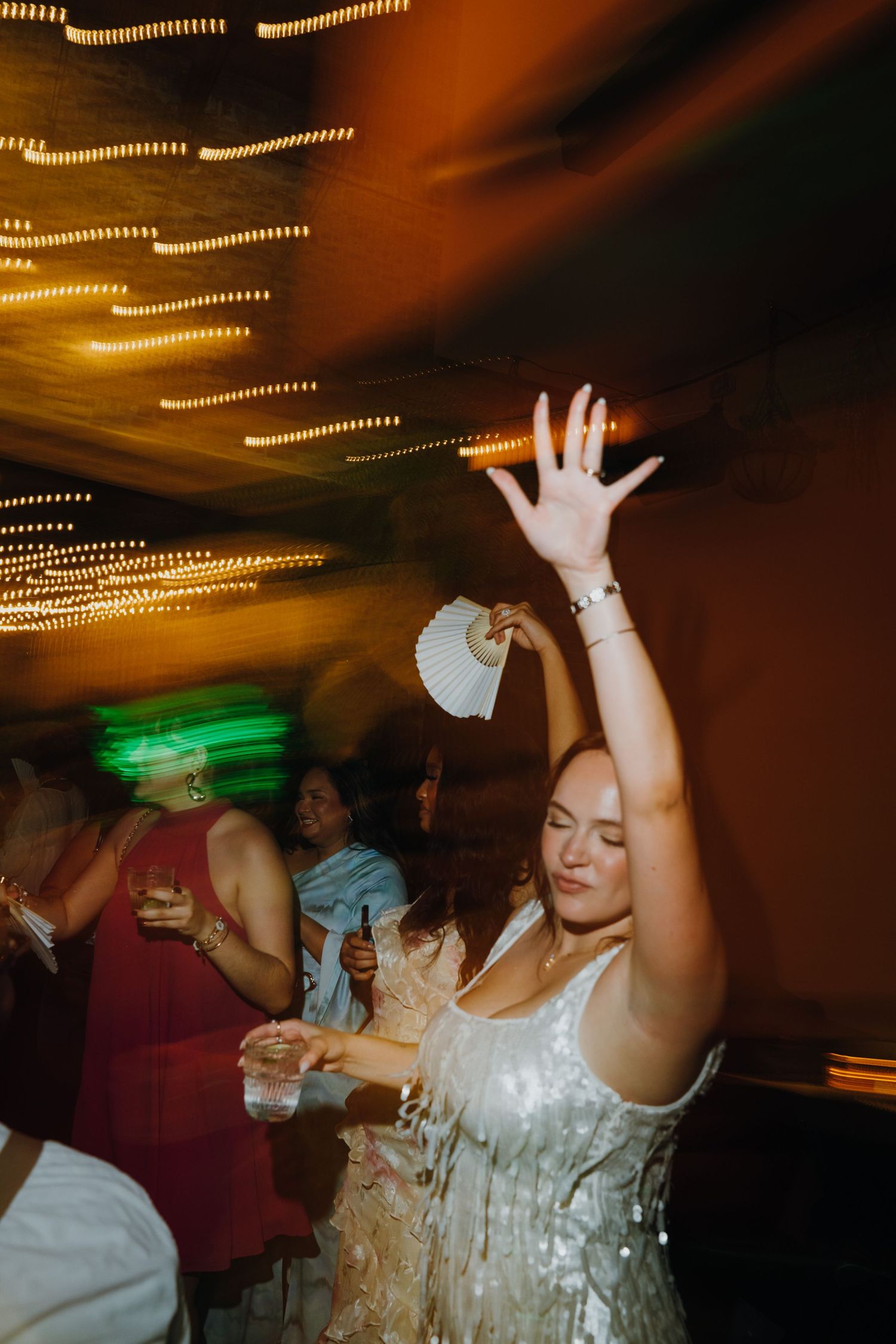 Bride in sparkly white dress dances with hand raised at energetic wedding reception under warm glowing lights. New Orleans, Louisiana, Missi