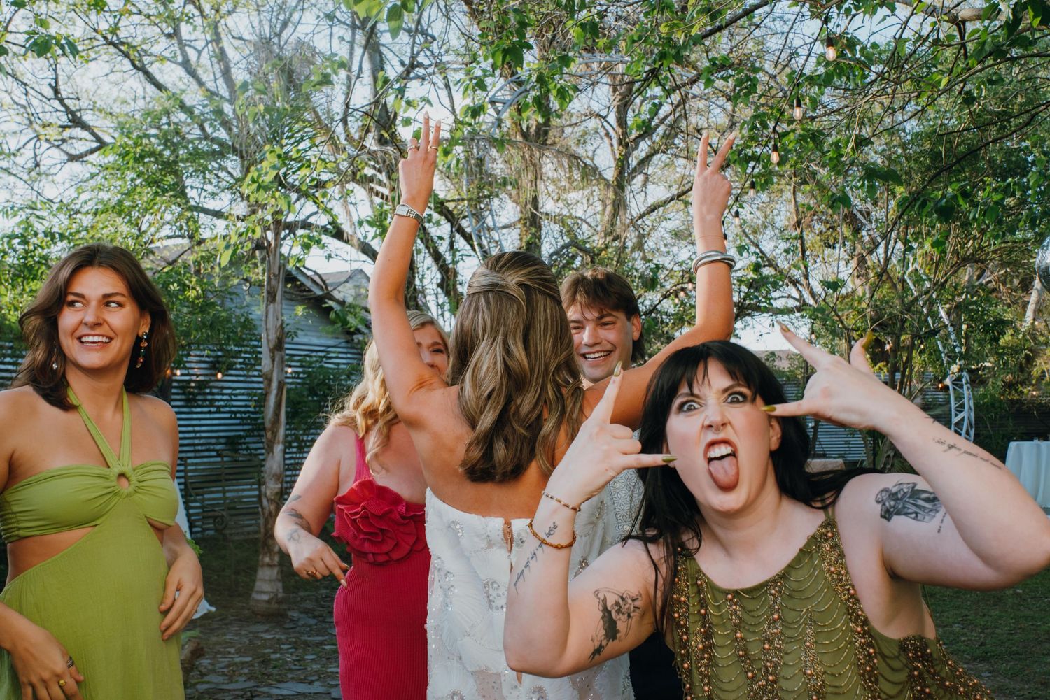 Group of joyful wedding guests dancing and celebrating outdoors under trees at a garden reception.