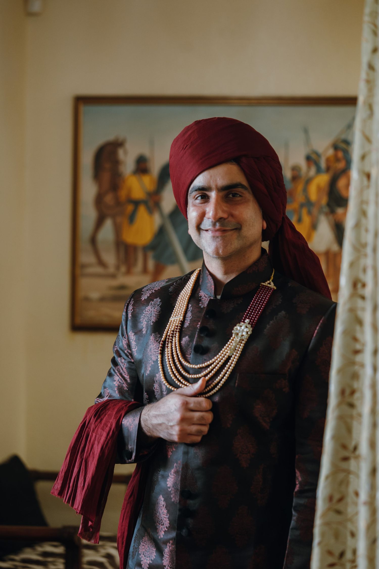 Handsome Indian groom in dark embroidered sherwani with layered pearl necklace and maroon turban posing.