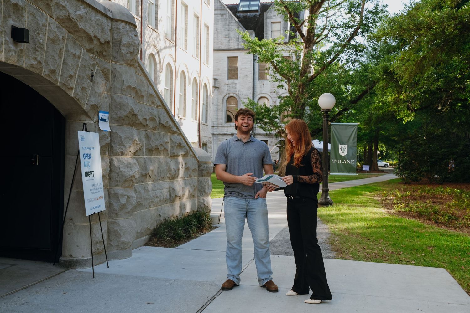 Two people standing outside a stone building on Tulane University campus reviewing documents near a green lamppost.