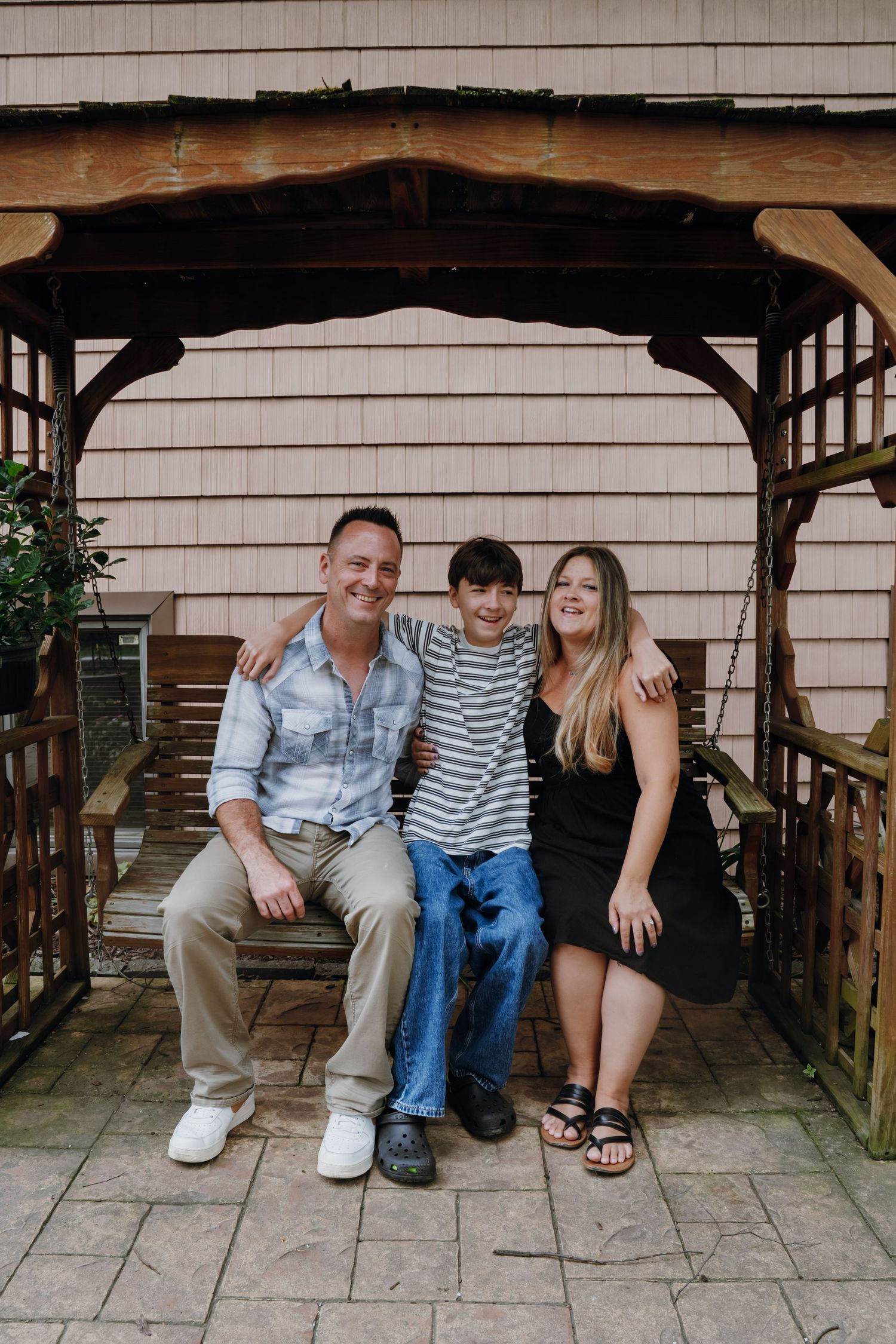 A happy family of three smiles together on a wooden swing under a pergola outside a beige home.