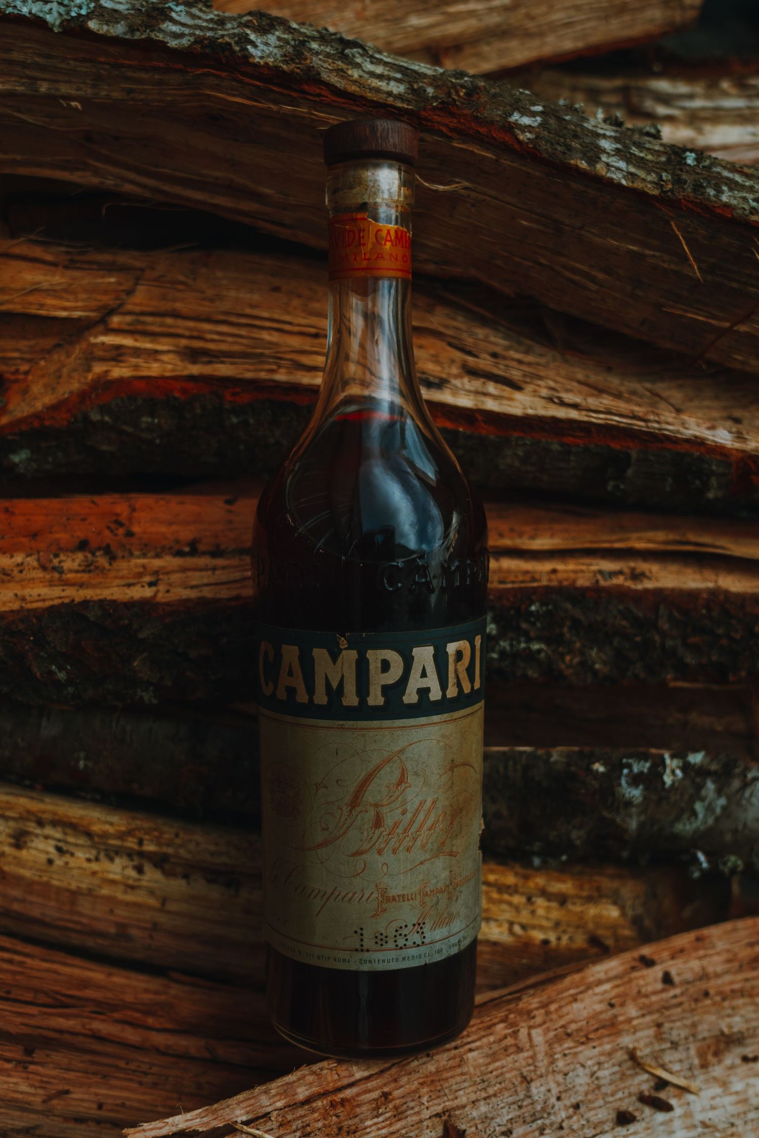 Vintage Campari bottle from 1963 resting against rustic weathered wooden logs.