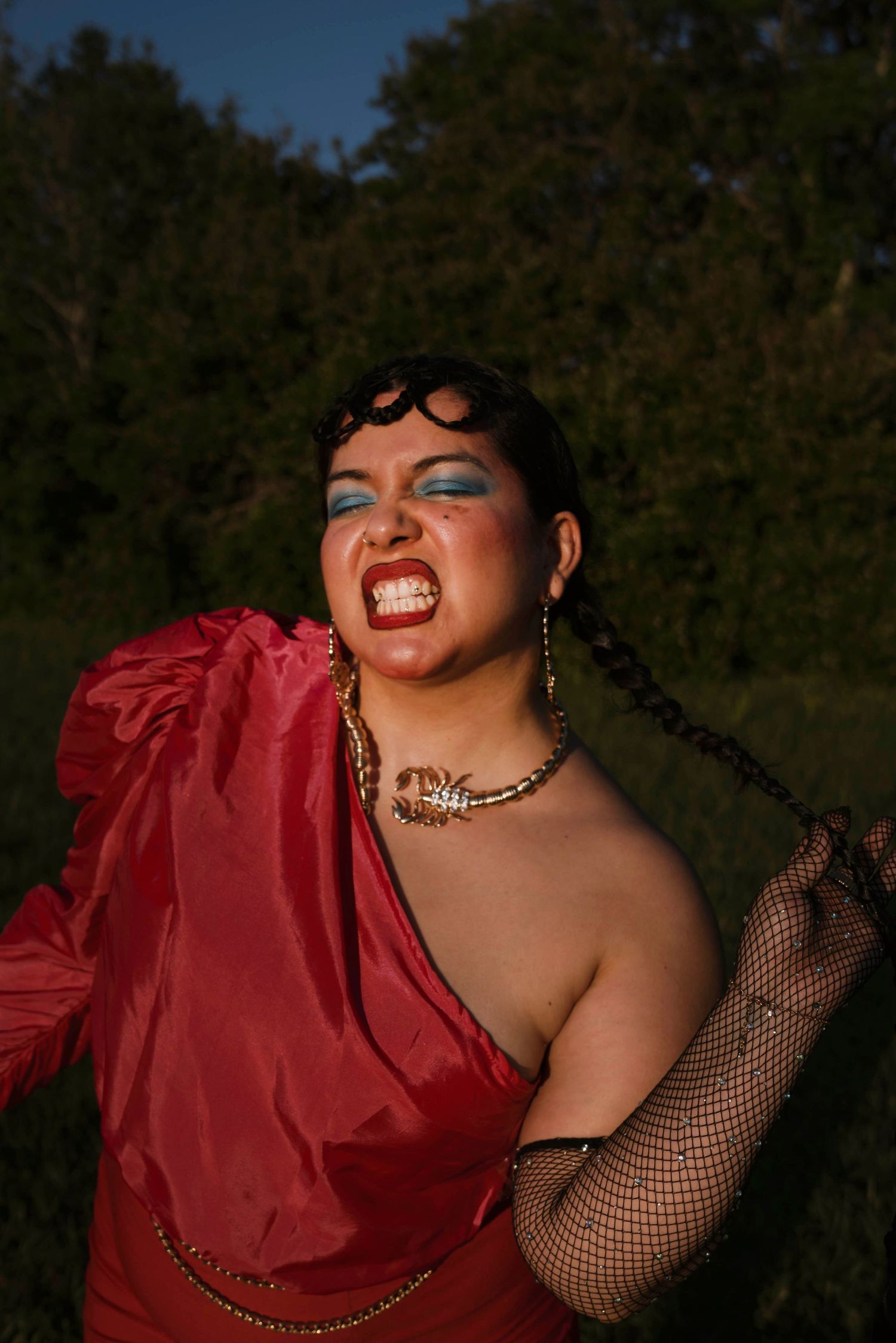 Woman in red dress with bold blue eyeshadow and scorpion jewelry posing dramatically outdoors.