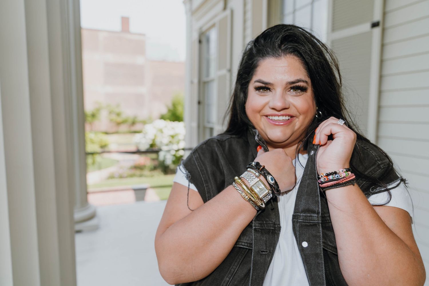 Smiling woman with bracelets and jewelry posing on a porch in a white top and dark vest.