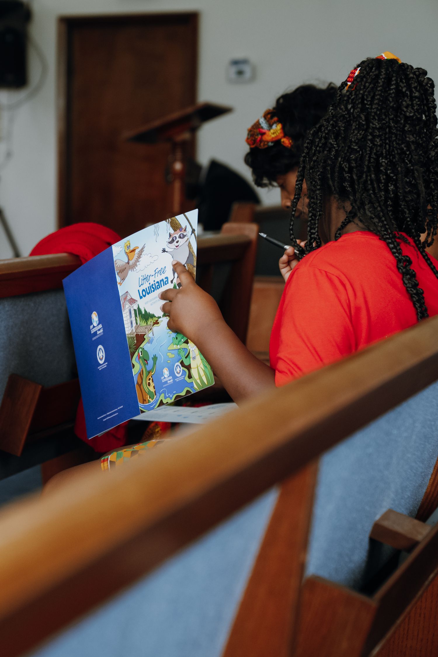 Young Black girl in red shirt reads Louisiana book while seated in church pew.
