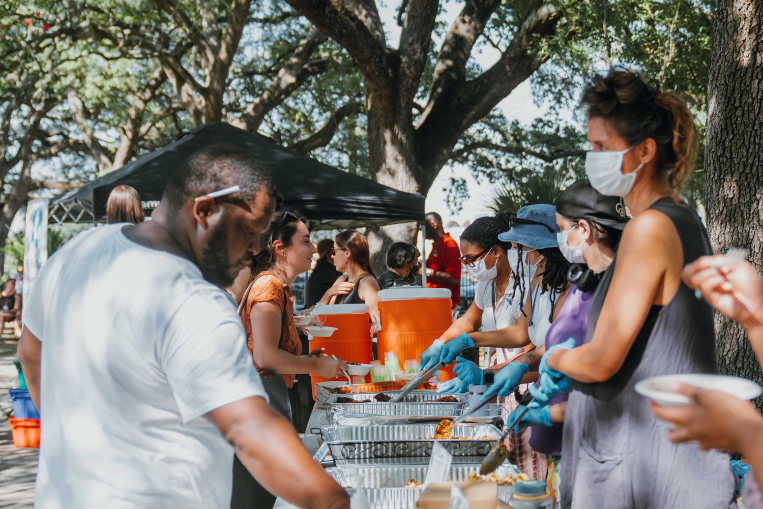 Volunteers in masks serve food to a crowd at an outdoor community event under shady trees.