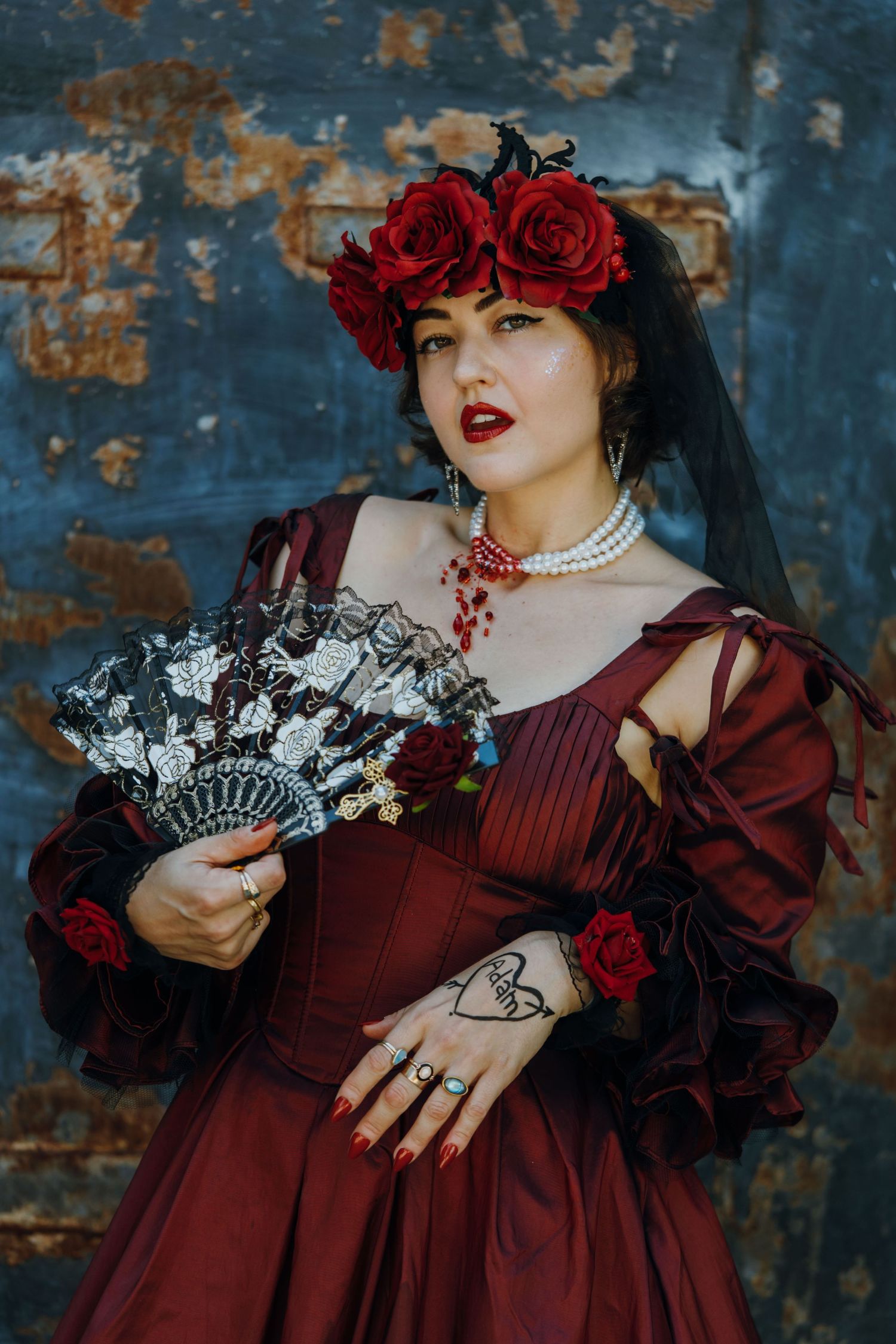 Woman in dark red gothic gown with rose crown, black veil, pearl necklace, and decorative fan poses dramatically.