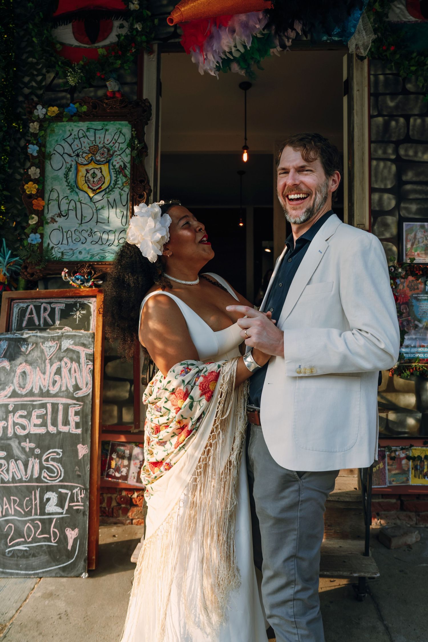 Laughing interracial couple in wedding attire stand outside a colorful art shop storefront.