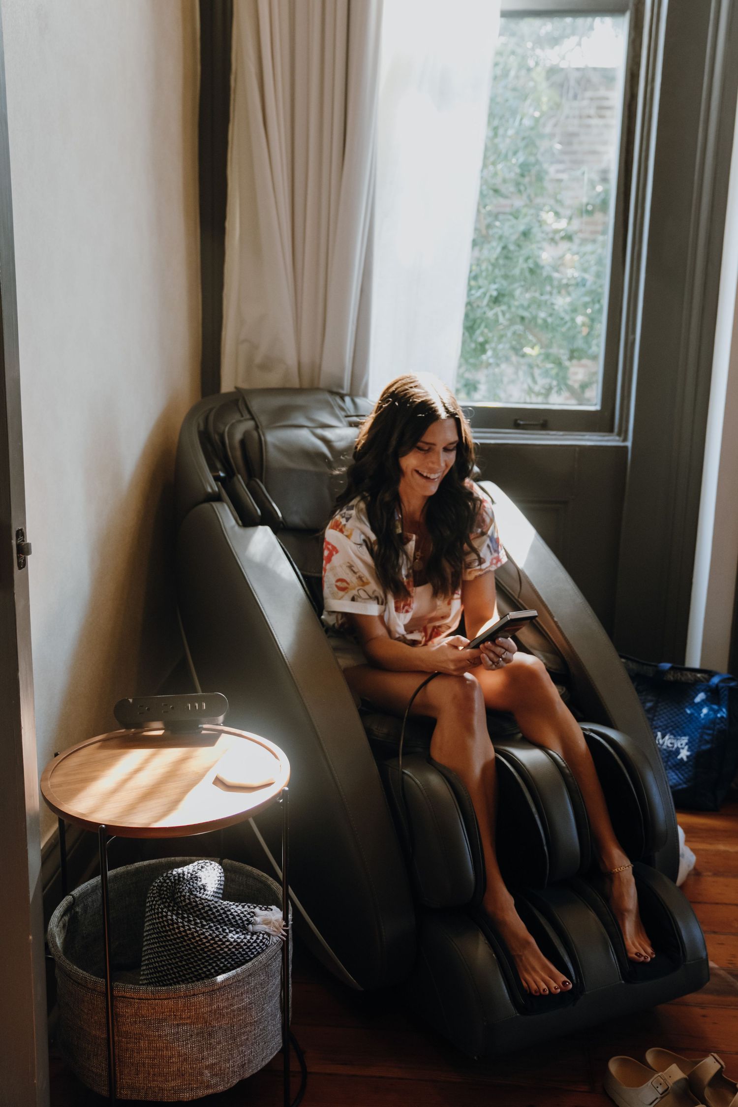Smiling woman relaxing in a black massage chair by a sunlit window on her phone.