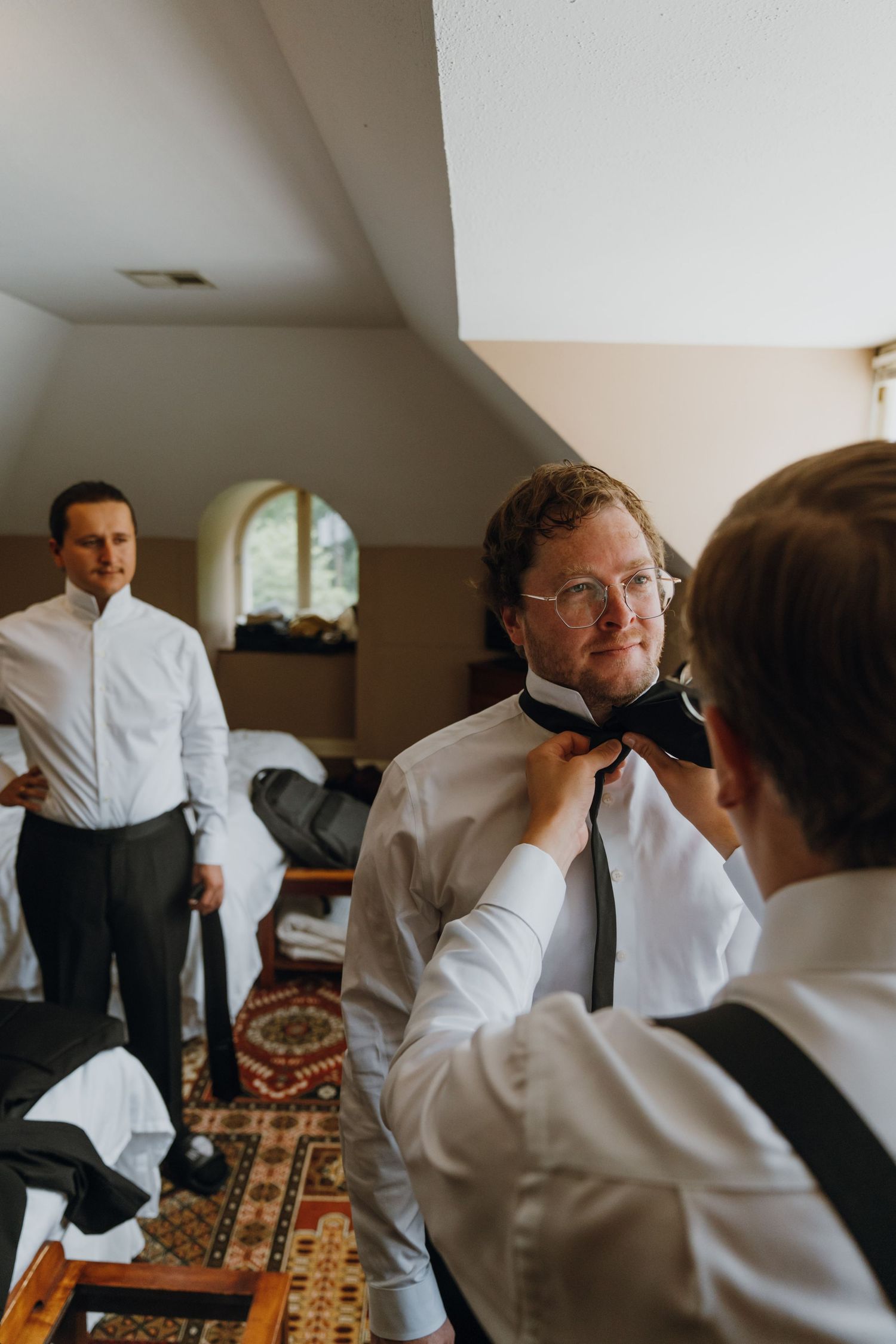 Groomsman helping groom adjust black bow tie while another groomsman dresses in background. New Orleans, Louisiana, Mississippi