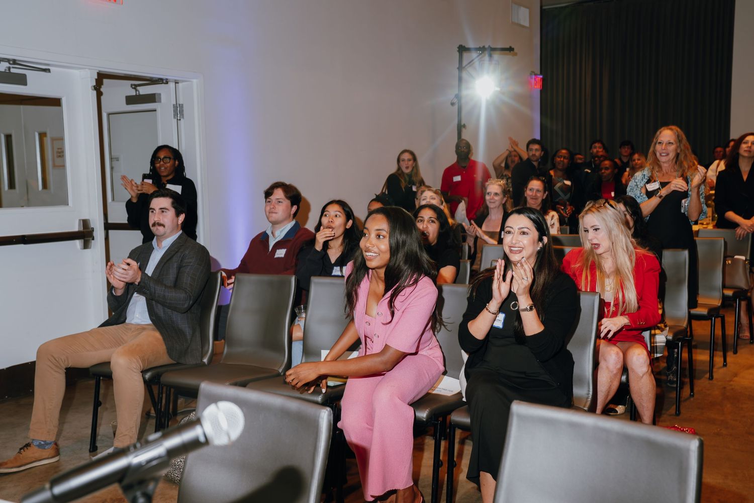 Diverse audience seated in chairs at an indoor event, engaged and applauding in a modern venue with bright lighting.