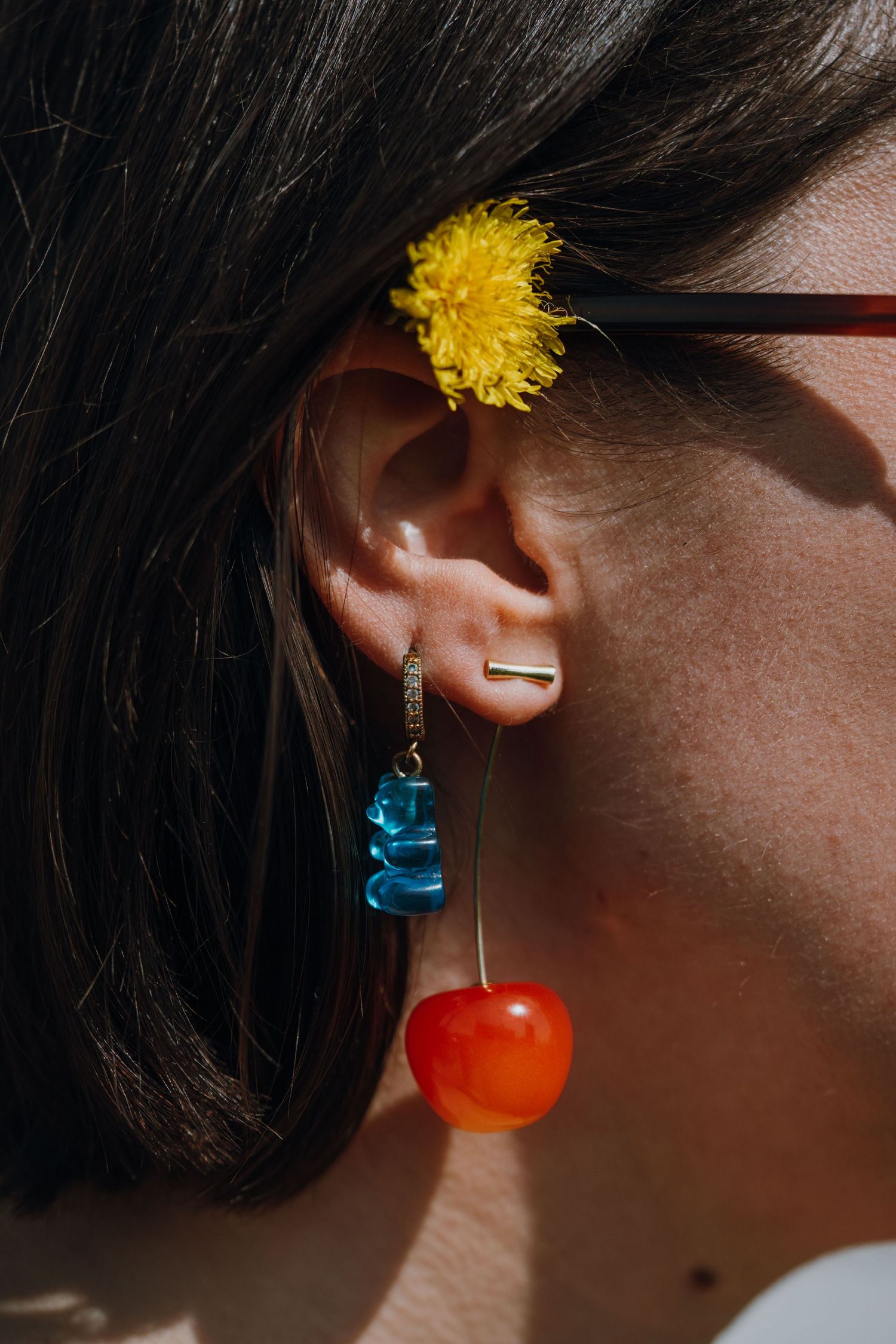 Close-up of woman's ear with blue gummy bear and cherry dangle earring and yellow flower.