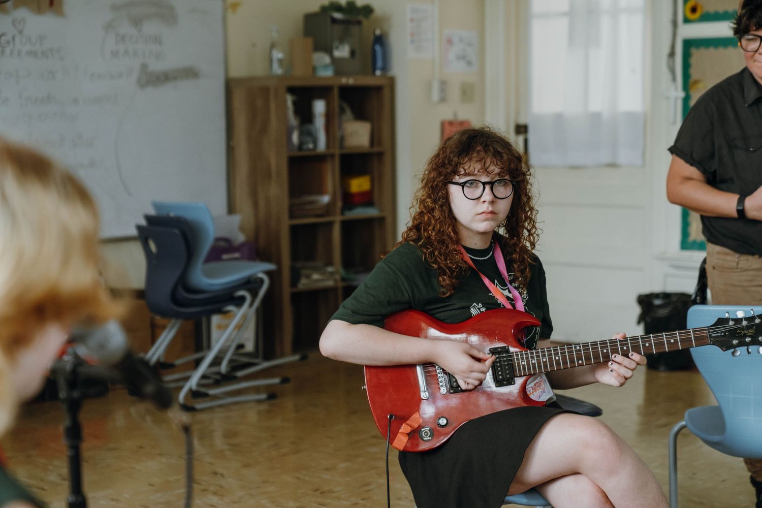 Curly-haired teen girl with glasses plays red electric guitar during music class session. New Orleans, Louisiana, Mississippi
