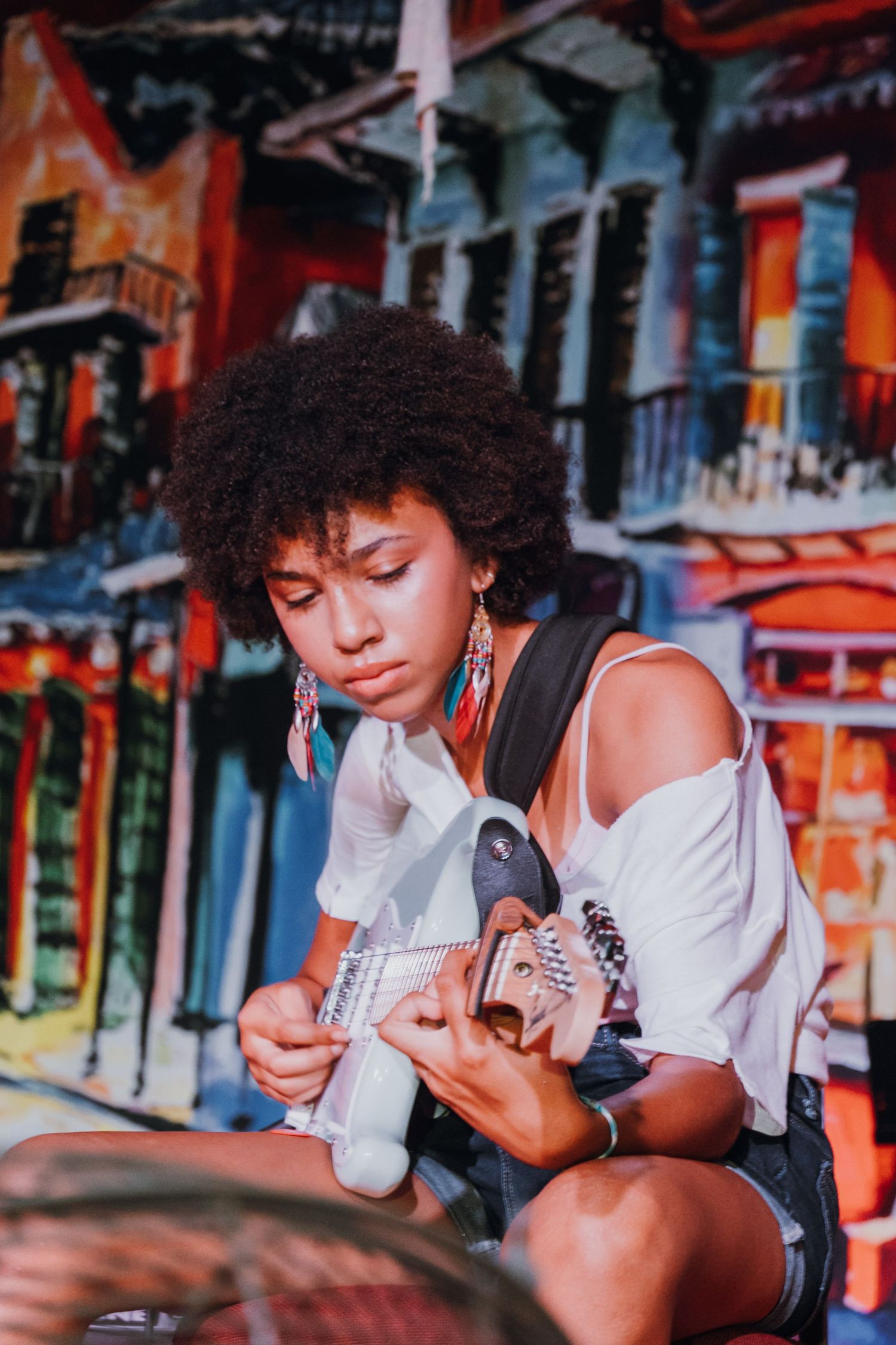 Young woman with afro hair playing electric guitar in front of a colorful New Orleans mural.