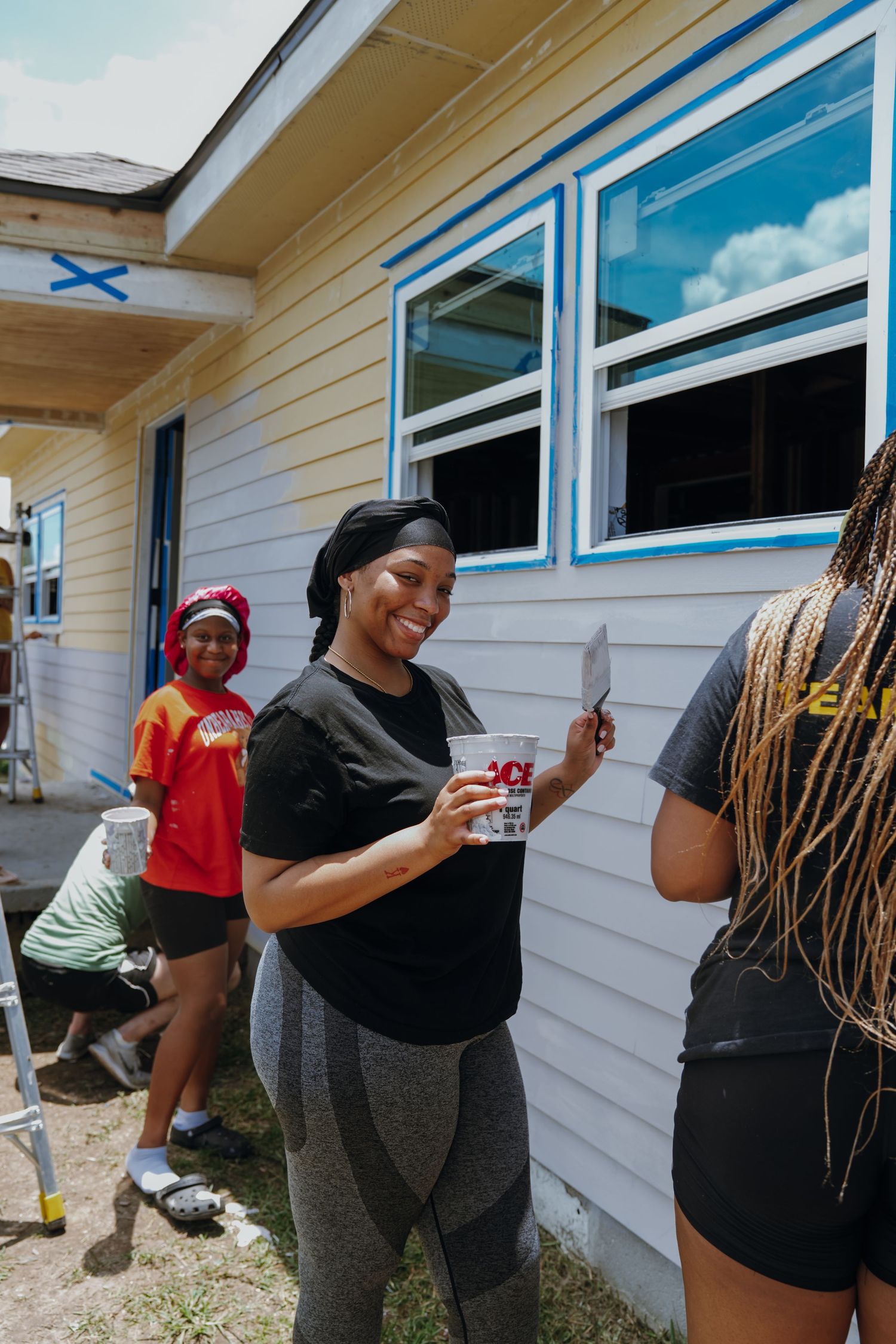 Smiling woman holding paint supplies while volunteering to paint a house exterior with other community volunteers.