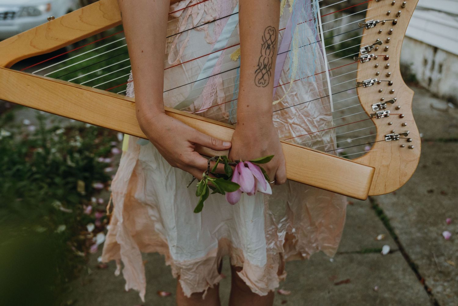 Close-up of woman in white dress holding a wooden harp decorated with pink flowers outdoors.