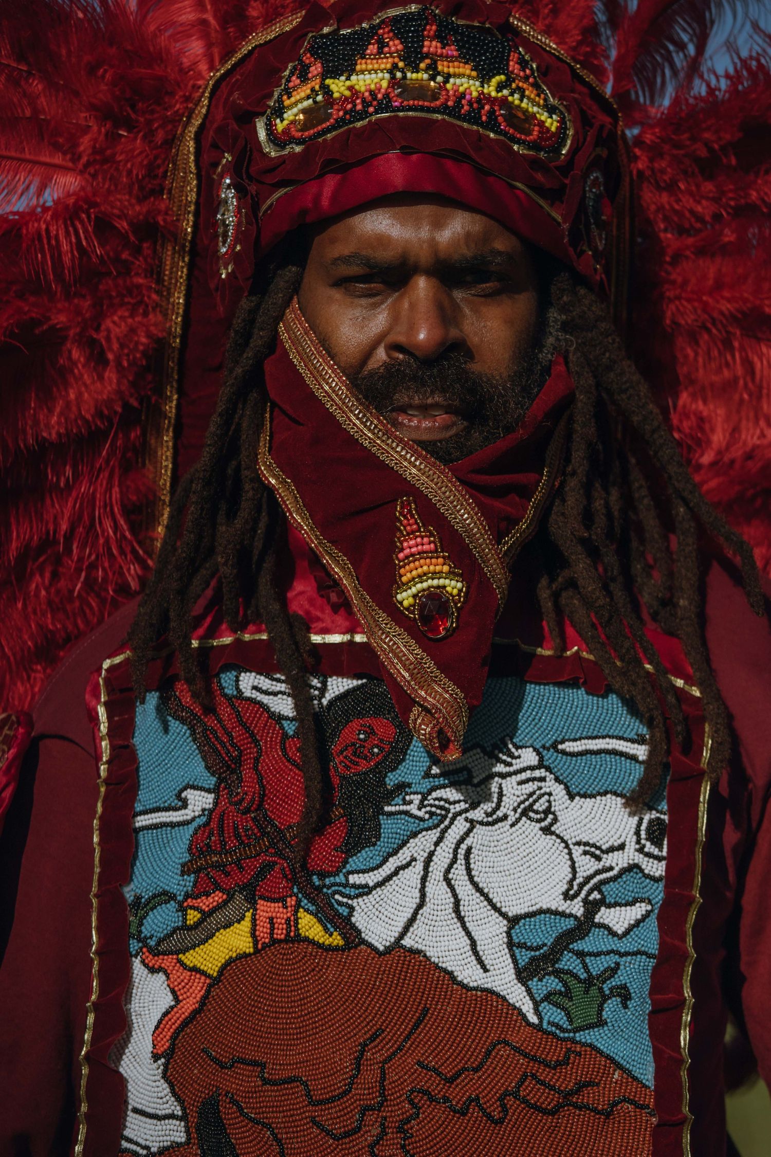 Portrait of a Mardi Gras Indian wearing a red beaded crown and feathered headdress with dreadlocks.