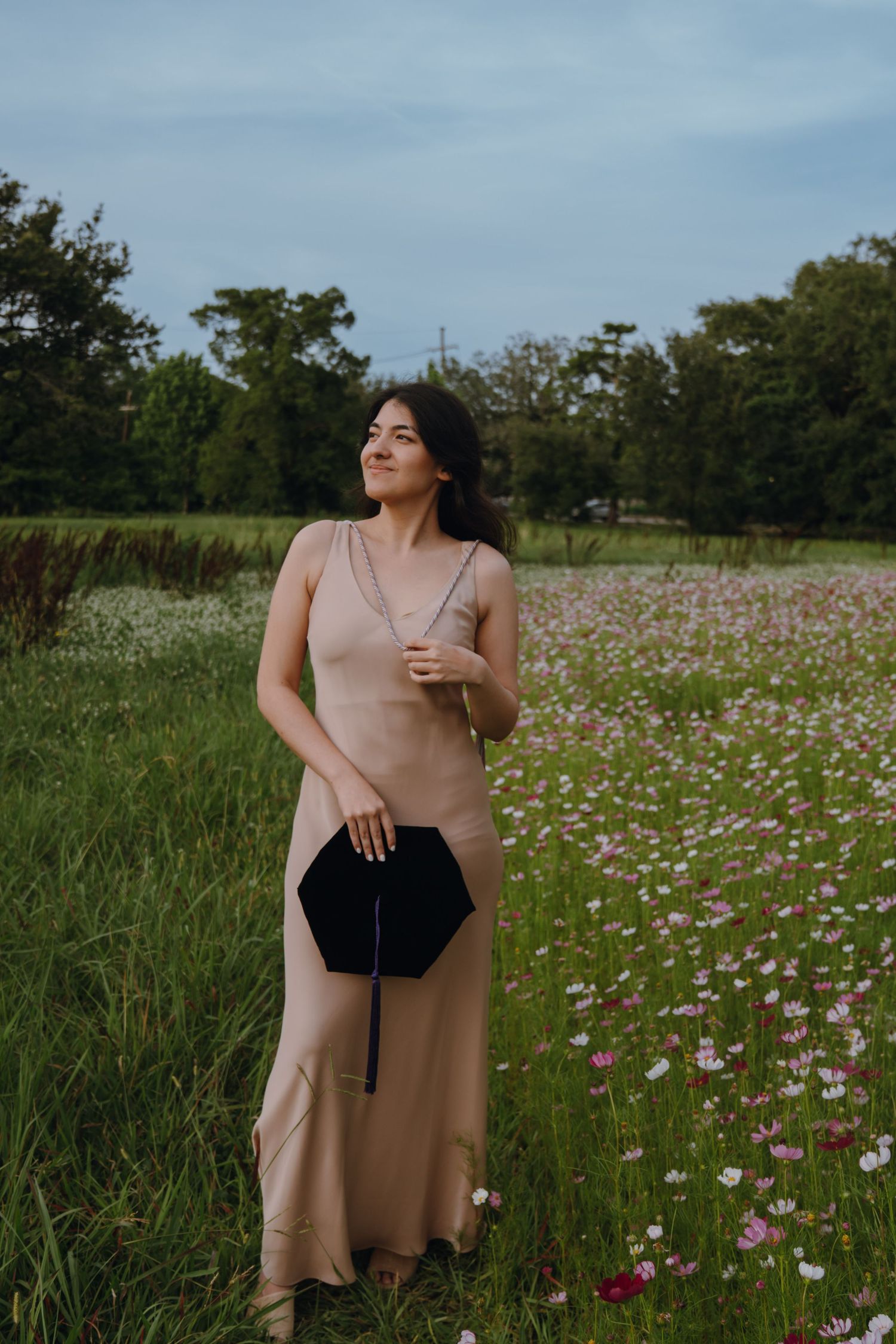 Young woman in elegant champagne slip dress holding black clutch in blooming flower field.