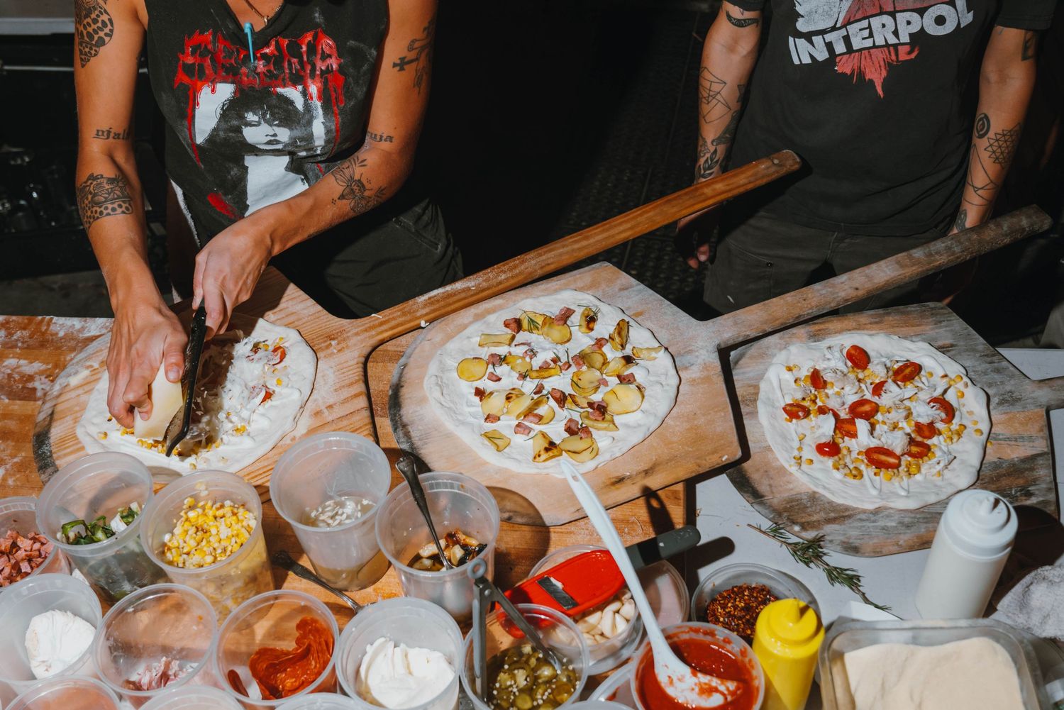 Tattooed chefs preparing multiple pizzas with colorful toppings on wooden boards at a pizzeria.